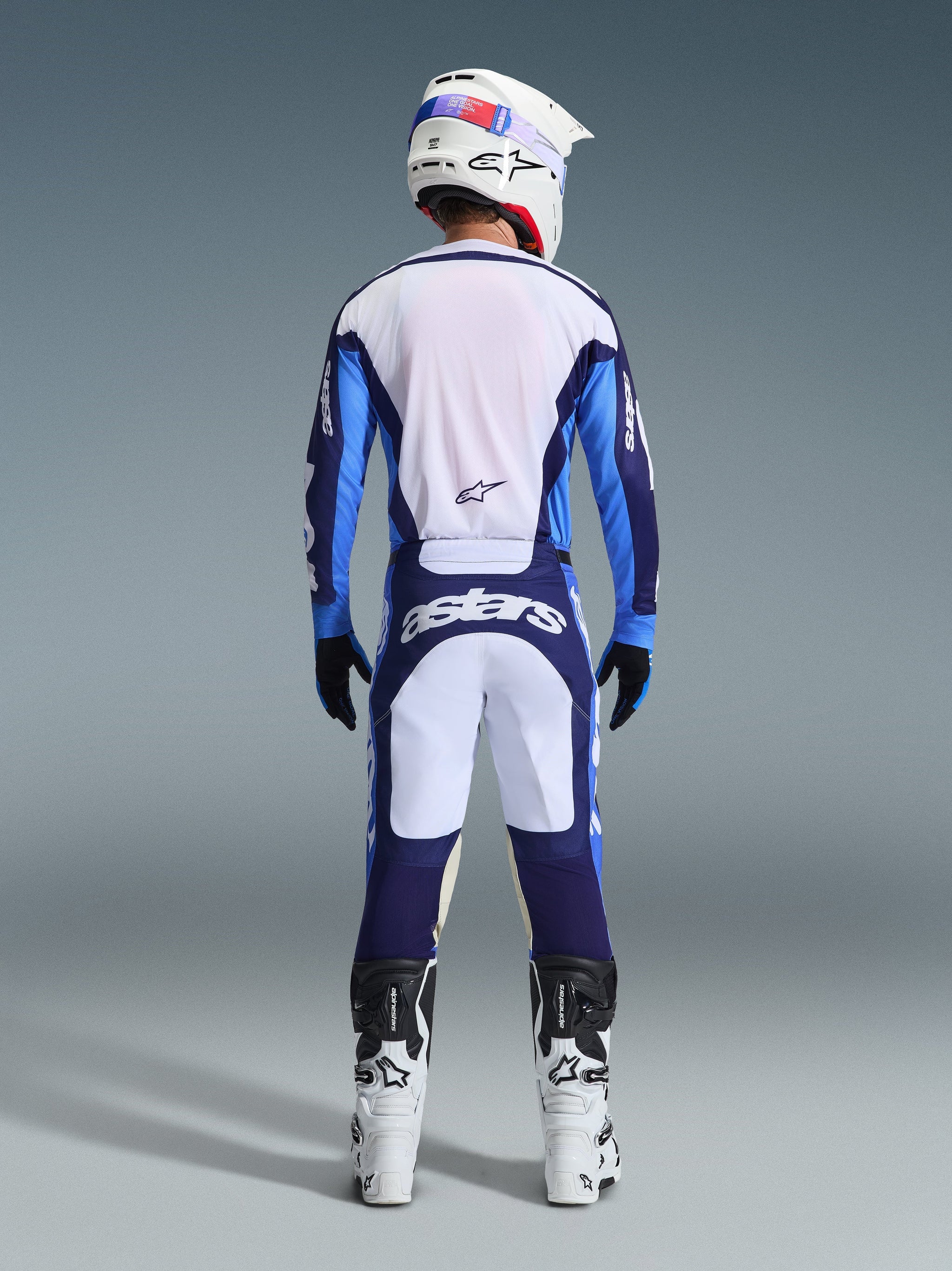 Motocross rider from behind wearing the Alpinestars 2026 Racer Riway Jersey in Ucla Blue and White, matching motocross pants with white and navy blue panels and Astars branding, white and black boots, and a white helmet, complete coordinated racing outfit for off-road performance.