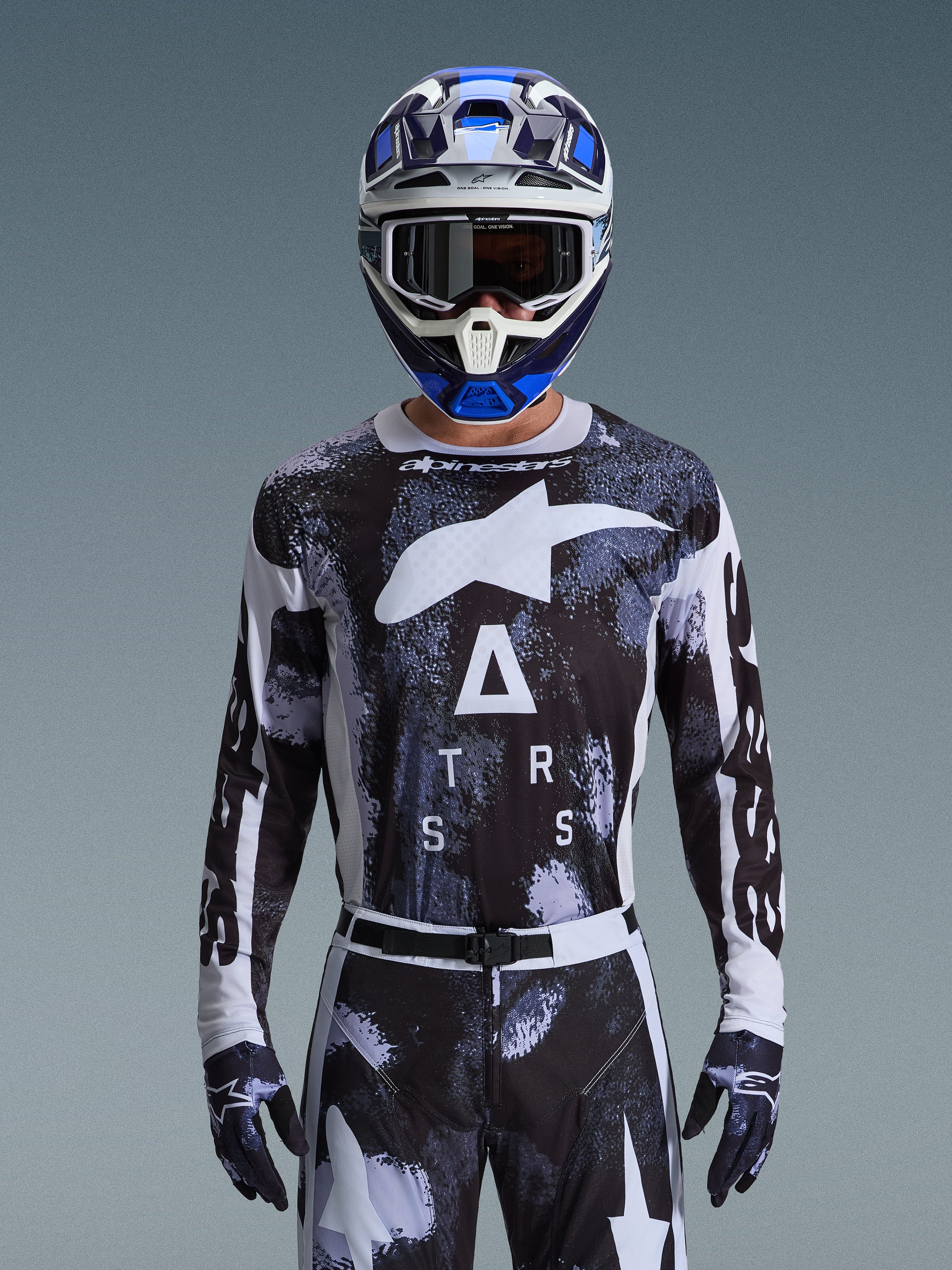 Motocross rider wearing the Alpinestars 2026 Racer Lahnd Gear Set, black and white abstract camo jersey with large white logos and "ATRS" lettering, matching pants and gloves, blue and white helmet with goggles, high-performance racing outfit for off-road motocross