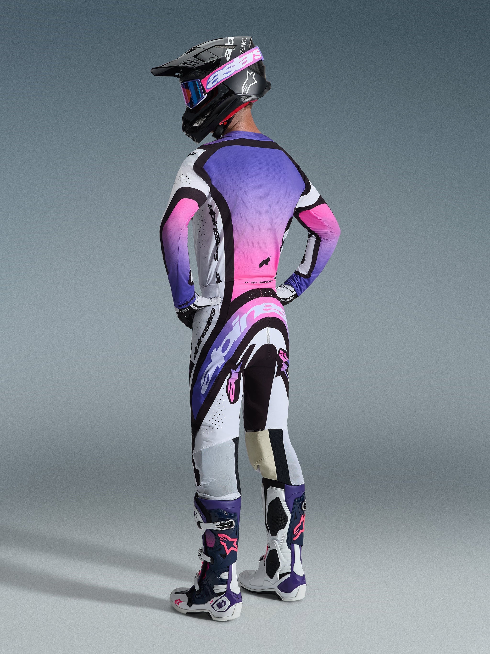 Alpinestars 2026 Supertech AFD Vista Jersey, Motocross Jersey, White Purple and Black with pink gradient, worn from behind with matching motocross pants, black helmet, and white/purple boots, showing the full professional racing gear set for off-road performance.
