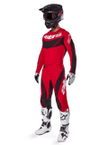 Full-body view of a motocross rider in the 2025 Techstar Dreem Gear Set in Bright Red and Black, featuring a performance jersey with white branding, matching stretch ripstop pants, a black helmet with a red visor, white and black gloves, and white Tech boots, specialized racing outfit for off-road competition.