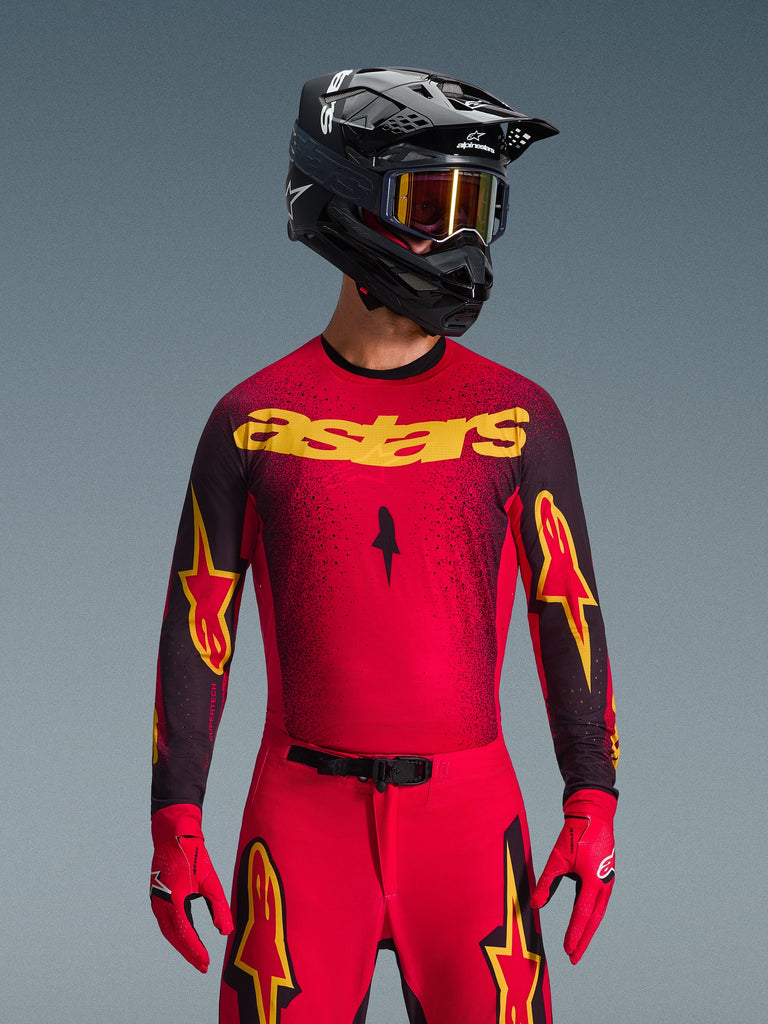 Alpinestars 2026 Supertech Scenz Jersey, Motocross Jersey, Red Black and Yellow, worn by a rider with a matching black helmet and red gloves, featuring a bold yellow 'astars' logo on the chest and a black gradient speckled pattern across the shoulders.
