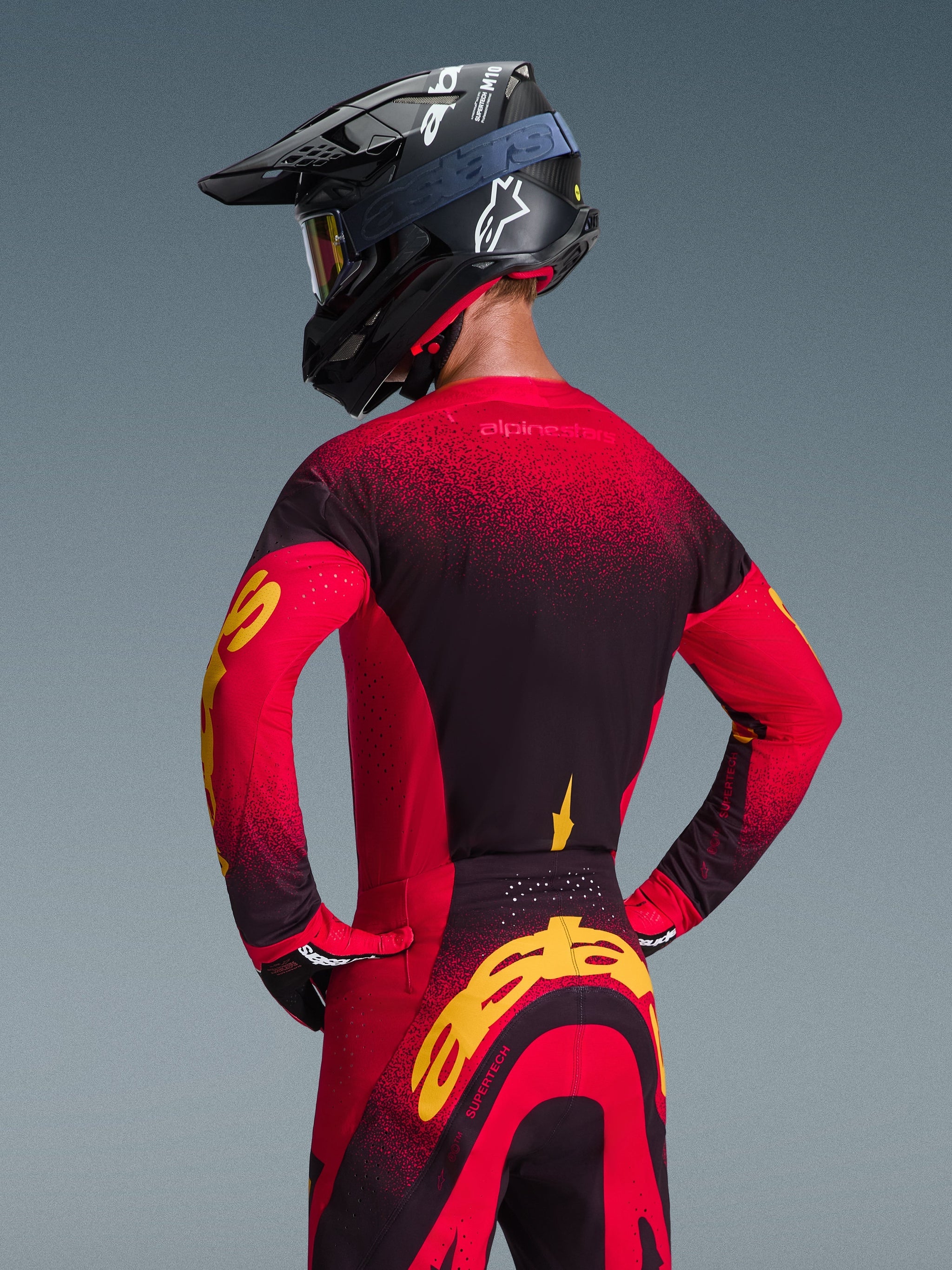 Alpinestars 2026 Supertech Scenz Jersey, Red Black and Yellow, back view of motocross rider wearing matching racing gear including off-road pants and a black helmet, featuring a red and black gradient design with bold yellow Alpinestars branding