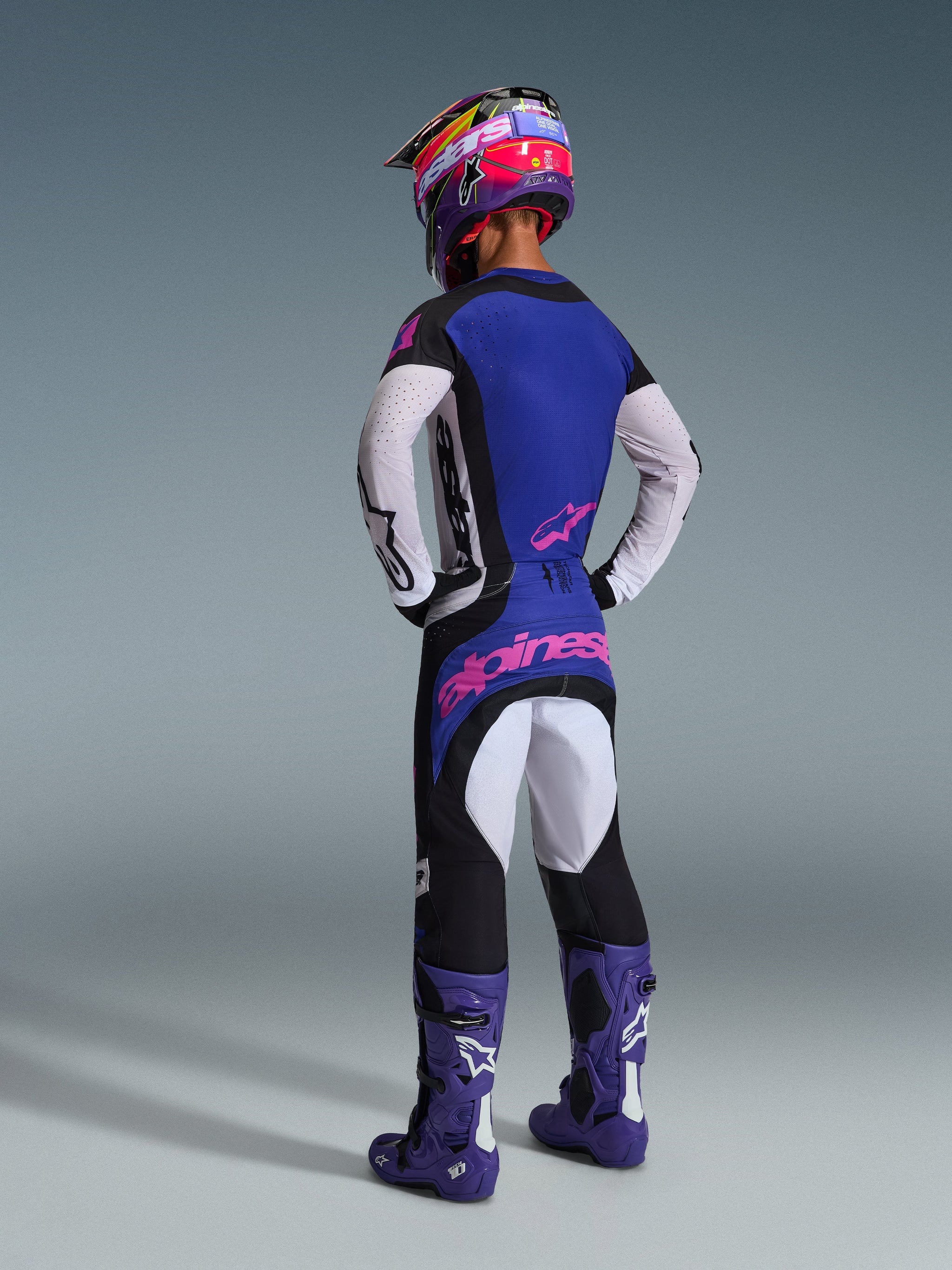Motocross rider from behind wearing complete racing gear, featuring the 2026 Techstar Knif Jersey with a blue back and white perforated sleeves, matching black pants with pink Alpinestars branding, colorful helmet, and purple boots, coordinated outfit for off-road competition.