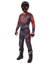 Motocross rider wearing complete racing gear, Alpinestars 2025 Racer Hollow Jersey and Pants in Gray Camo Orange Fluo, black helmet with orange goggles, black and red gloves, and black boots with white logos, coordinated performance outfit for off-road racing