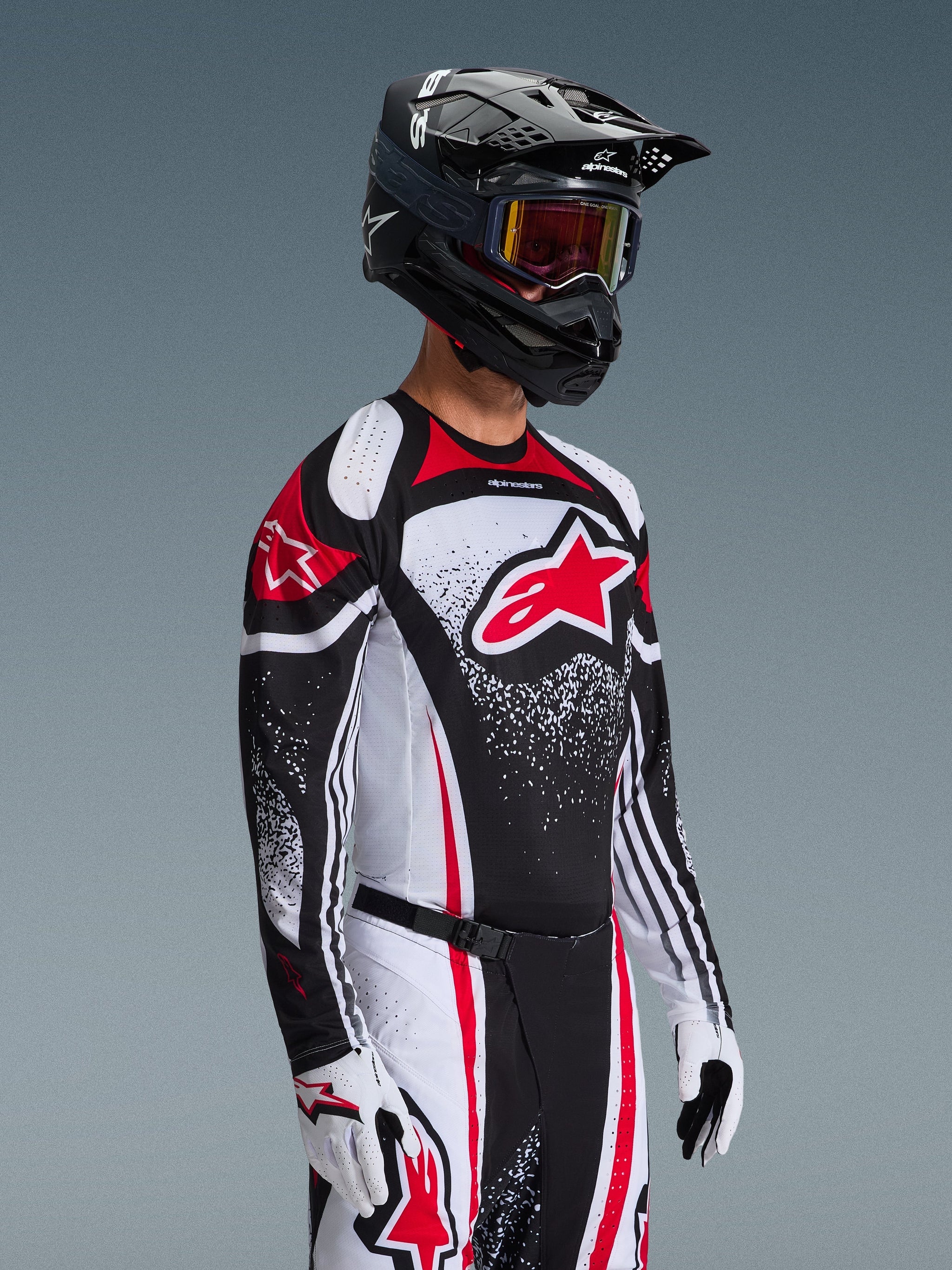 Motocross rider wearing the Alpinestars 2026 Techstar Nomur Jersey in Black White Red, featuring a large red Astars logo and speckled graphic patterns, paired with matching racing pants, white gloves, and a black helmet with goggles, coordinated outfit for off-road competition