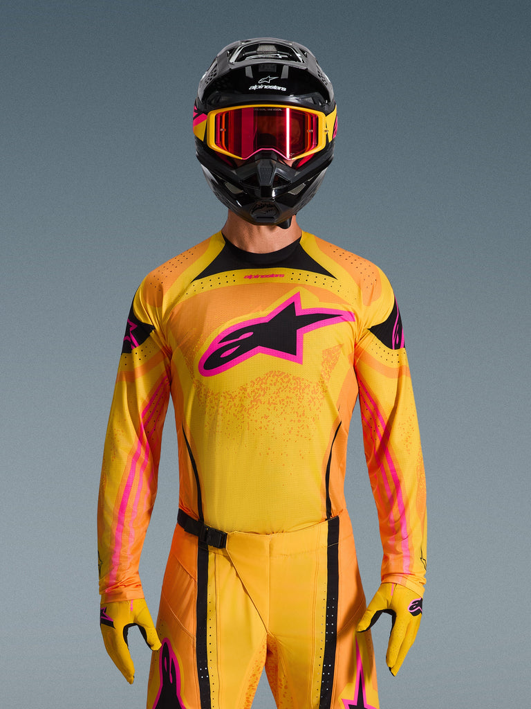 2026 Techstar Nomur Jersey in Orange Yellow Pink, worn by a motocross rider with a matching black helmet and pink goggles, featuring a large pink Alpinestars logo on the chest, athletic race fit for off-road performance.
