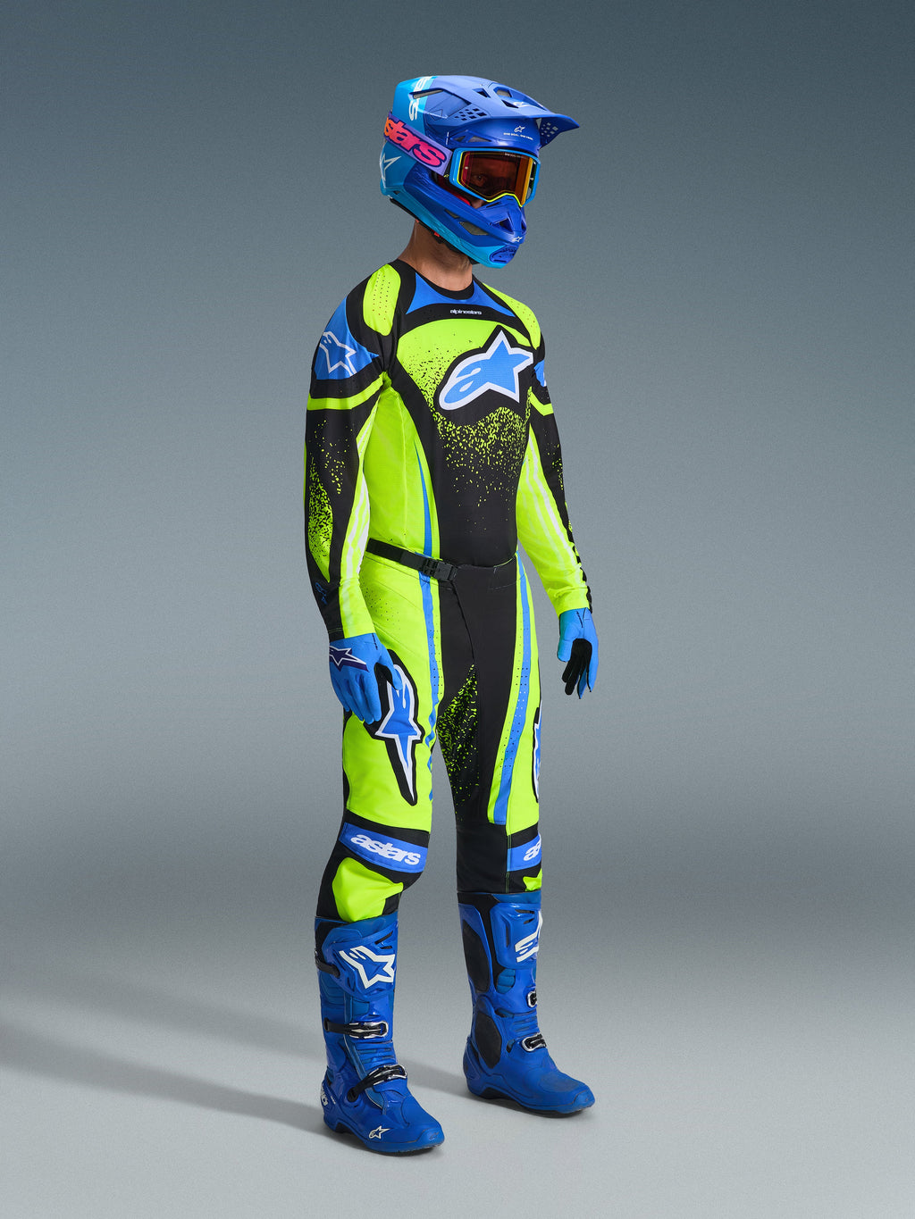 Motocross rider wearing the Alpinestars 2026 Techstar Nomur Gear Set, bright lime green jersey and pants with black panels and blue accents, large blue star logo on chest and thighs, blue helmet with orange goggles, blue boots, and blue gloves, full racing kit for motocross