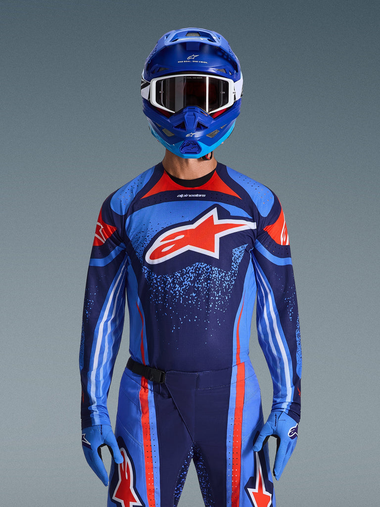 Motocross rider wearing the Alpinestars 2026 Techstar Nomur Jersey in Dark Navy, Orange, and UCLA Blue, matching pants and blue gloves, blue helmet with white goggles, large red and white Alpinestars logo on the chest, coordinated performance gear for off-road racing.