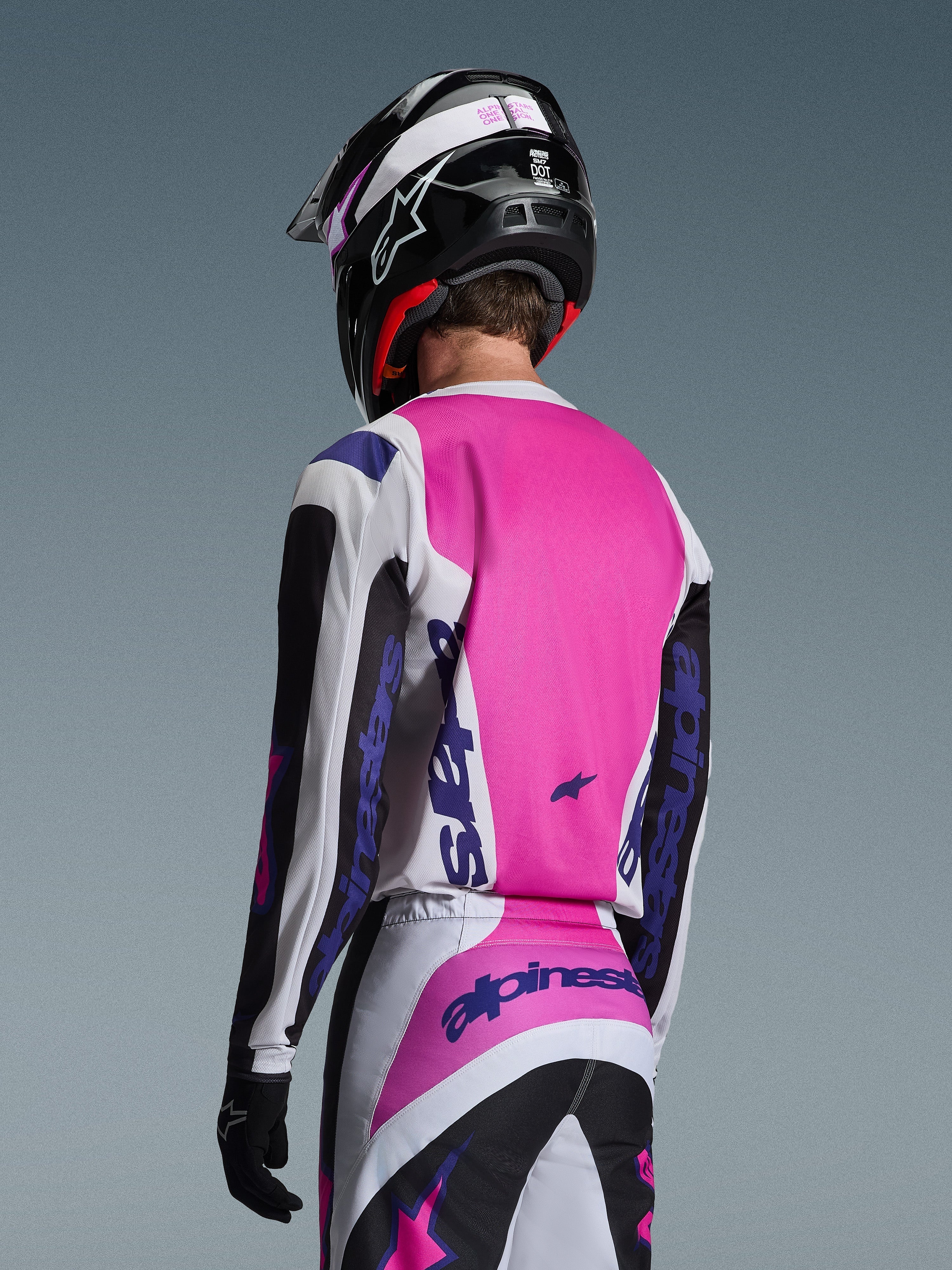 Motocross rider seen from the back wearing the Alpinestars 2026 Fluid Grid Jersey in Black, White, and Purple, matching pants with bold branding, black helmet with pink accents, and black gloves, complete coordinated gear for off-road performance.