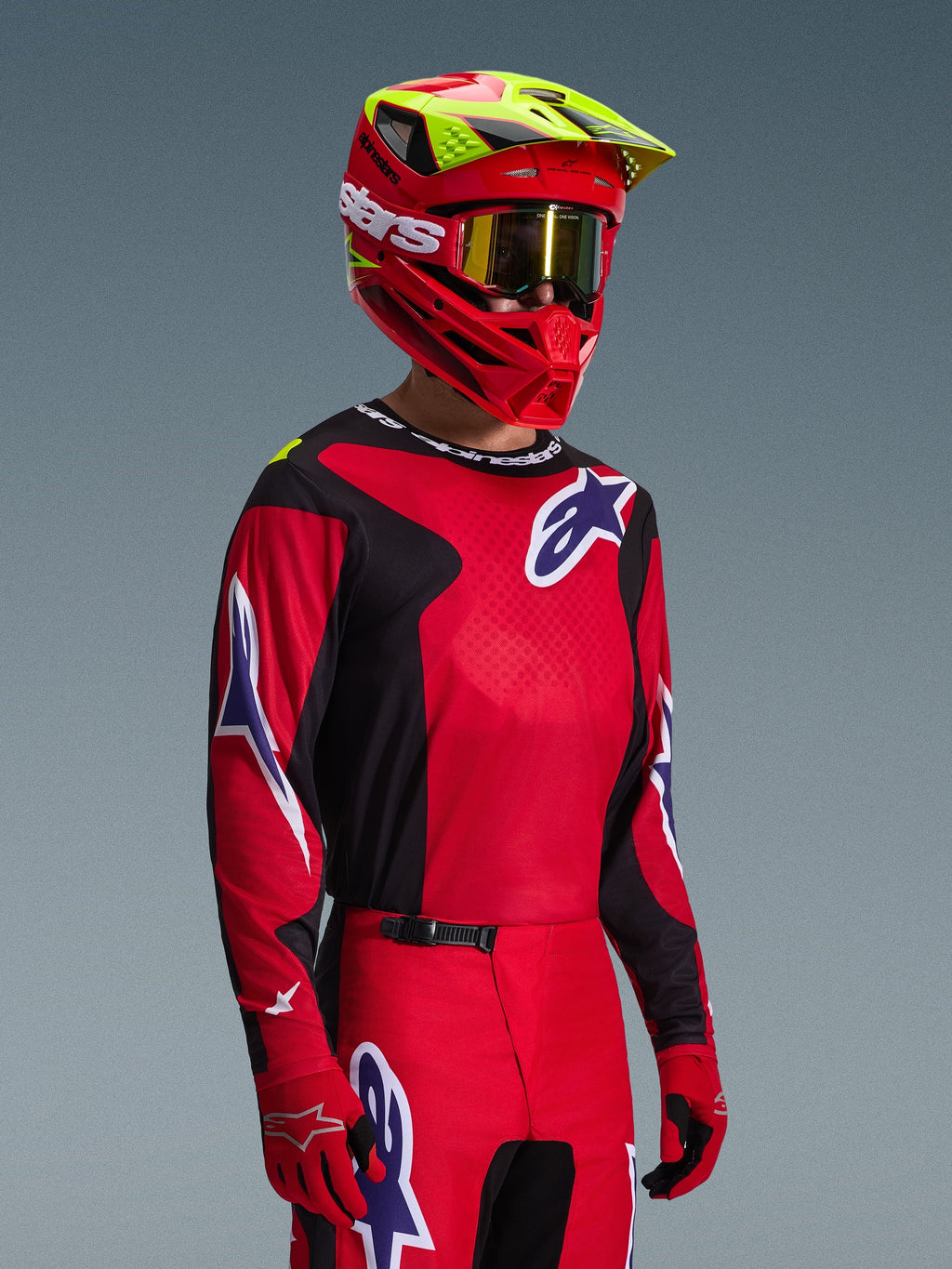 Motocross rider wearing the Alpinestars 2026 Fluid Grid Jersey in Red Black and Purple, coordinated with matching motocross pants, a red helmet with neon yellow accents, gold goggles, and red gloves, complete off-road racing outfit.