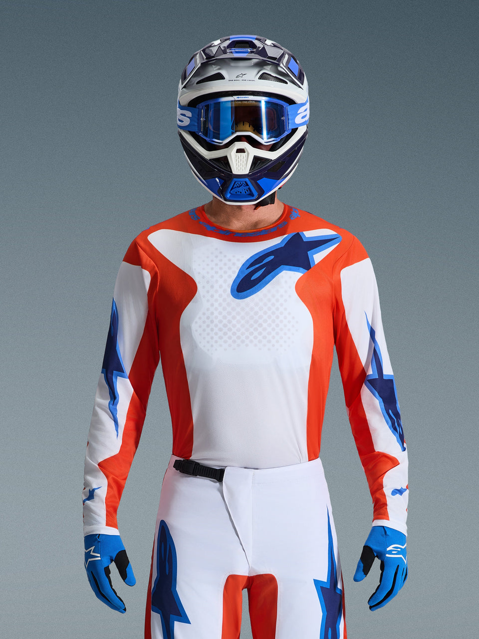 Motocross rider wearing the 2026 Fluid Grid Jersey in Orange Ucla Blue, featuring a white chest with orange side panels and blue Alpinestars logos, coordinated with matching white and blue pants, blue gloves, and a white and blue helmet for a professional off-road racing look.