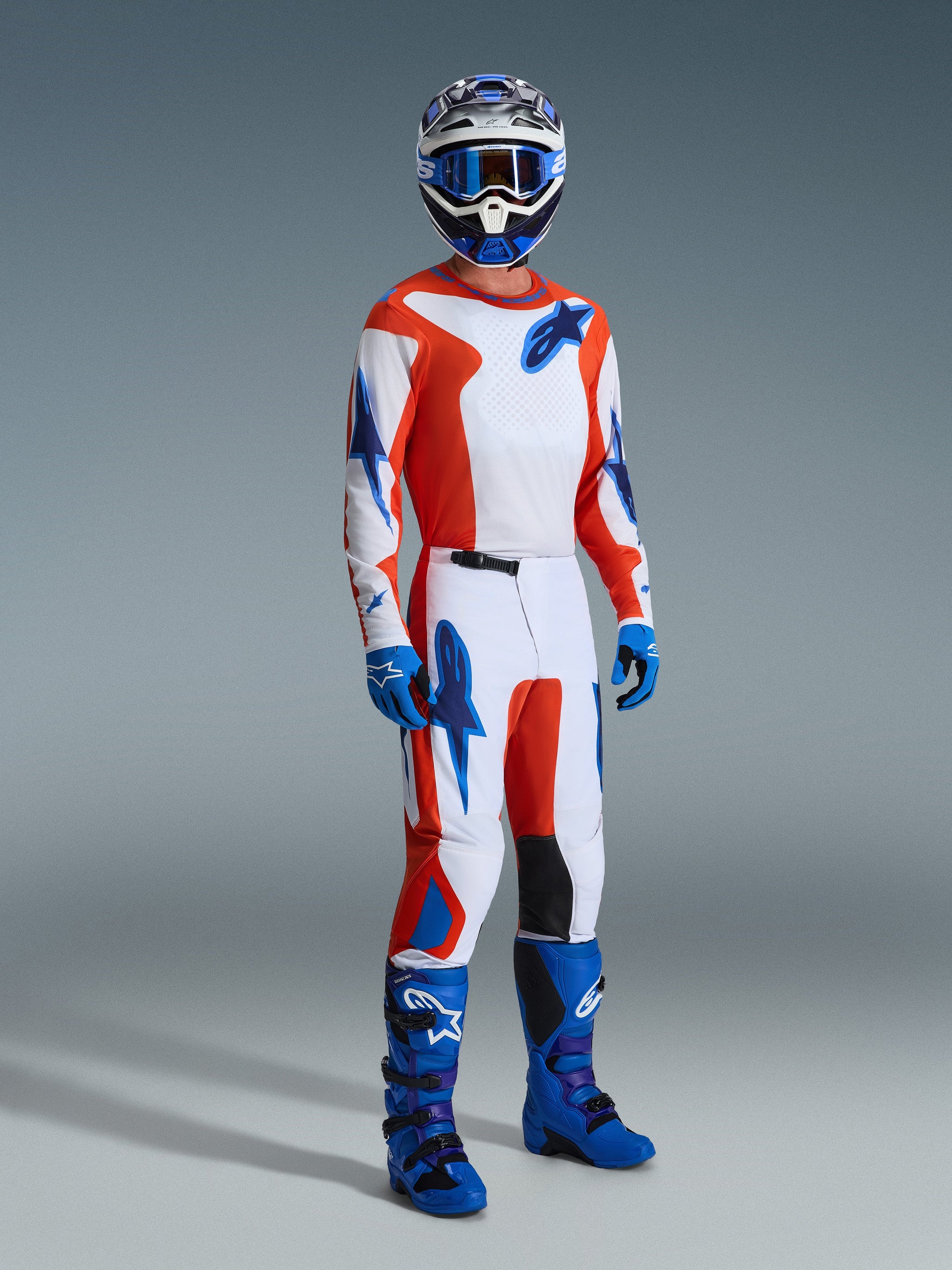 Motocross rider wearing complete racing gear, 2026 Fluid Grid Jersey in Orange and UCLA Blue, matching pants with white panels and blue logos, blue motocross boots, and a coordinated helmet, professional studio shot for off-road racing