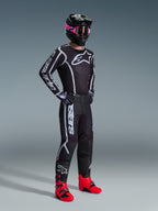 Alpinestars 2026 Fluid Apex Gear Set, motocross rider wearing a black jersey and pants with white and grey graphic accents, black helmet with pink goggles, and bright pink racing boots, coordinated professional motocross outfit