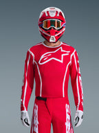 Motocross rider wearing Alpinestars 2026 Fluid Apex Gear Set, Red jersey with large white star logo and white piping, matching red pants with white accents, red and white helmet with purple highlights and orange-tinted goggles, white and black gloves, coordinated outfit for off-road racing
