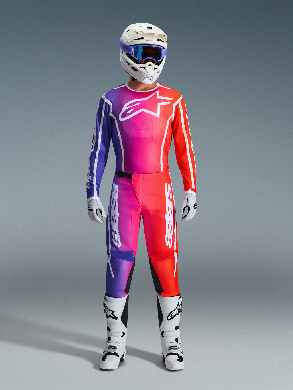 Motocross rider wearing a complete Alpinestars 2026 Fluid Apex racing gear set, featuring a purple, pink, and orange gradient jersey and pants. The coordinated outfit includes a white Alpinestars logo on the chest, a white helmet with blue goggles, and white boots, designed for off-road performance.