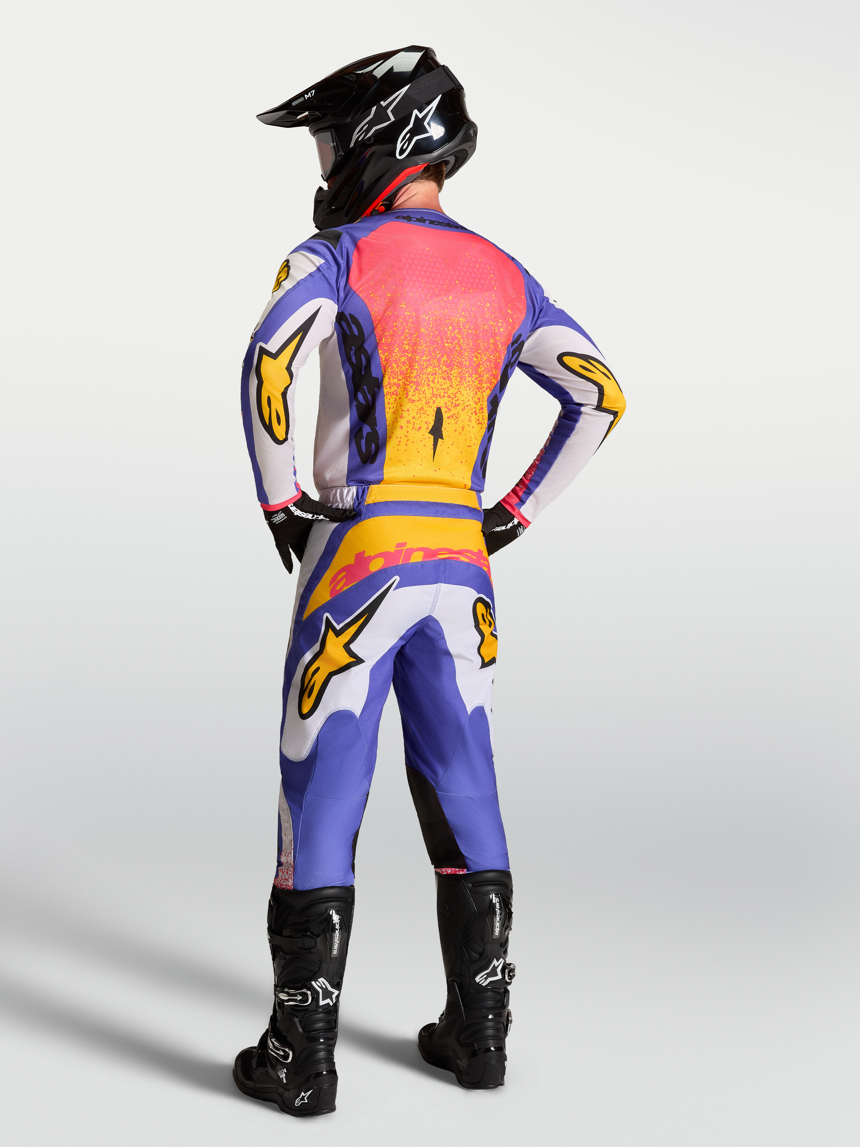 Alpinestars Fluid Drag Jersey in lilac and blue with gold accents, shown on a motocross rider from behind in full gear.