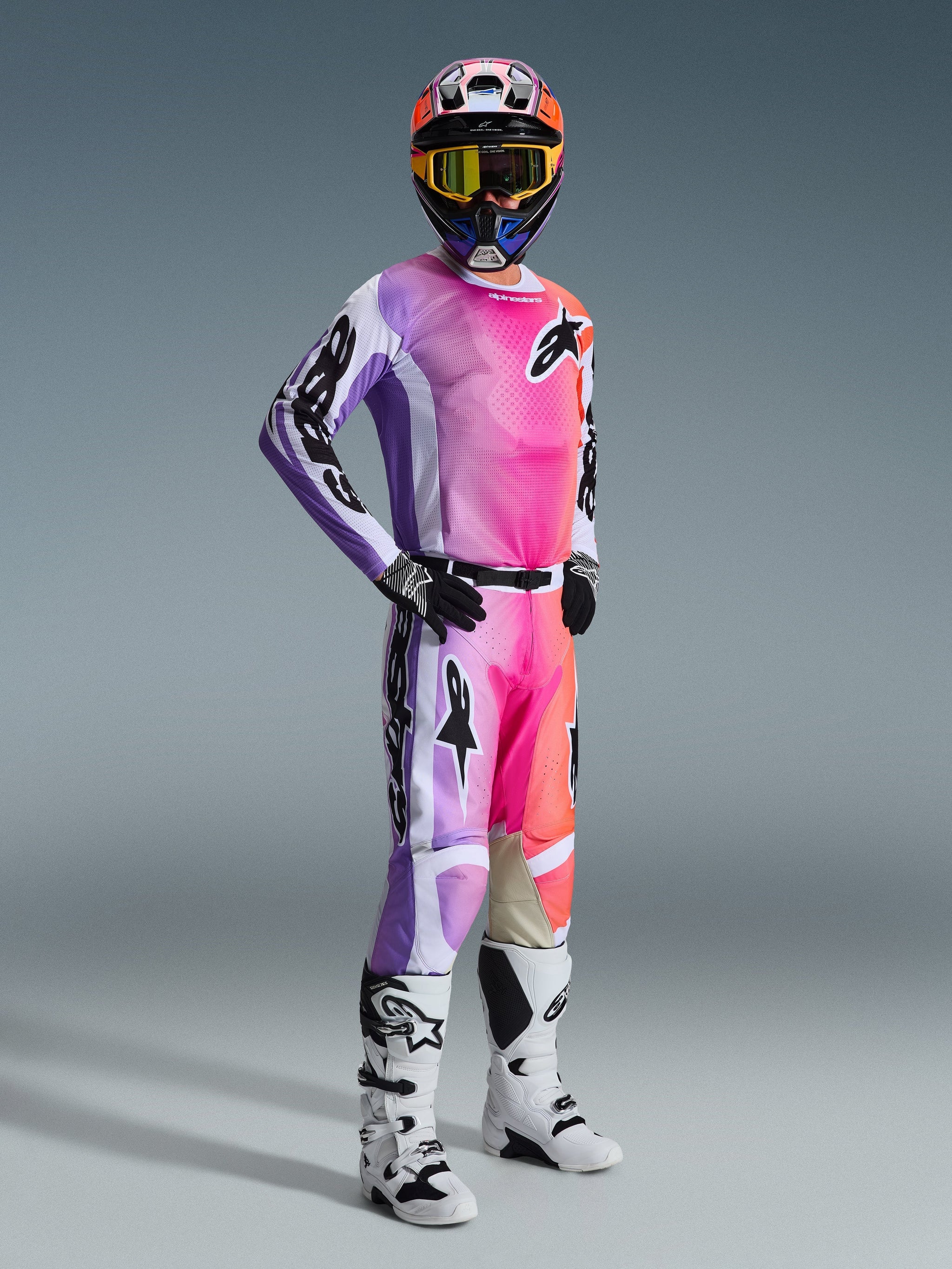 Motocross rider wearing complete racing gear featuring the Alpinestars 2026 Racer Air Portl Pants, vibrant pink, purple, and orange multicolor design with white accents, matching long-sleeve jersey, black helmet with yellow goggles, and white boots, coordinated outfit for high-performance off-road racing.