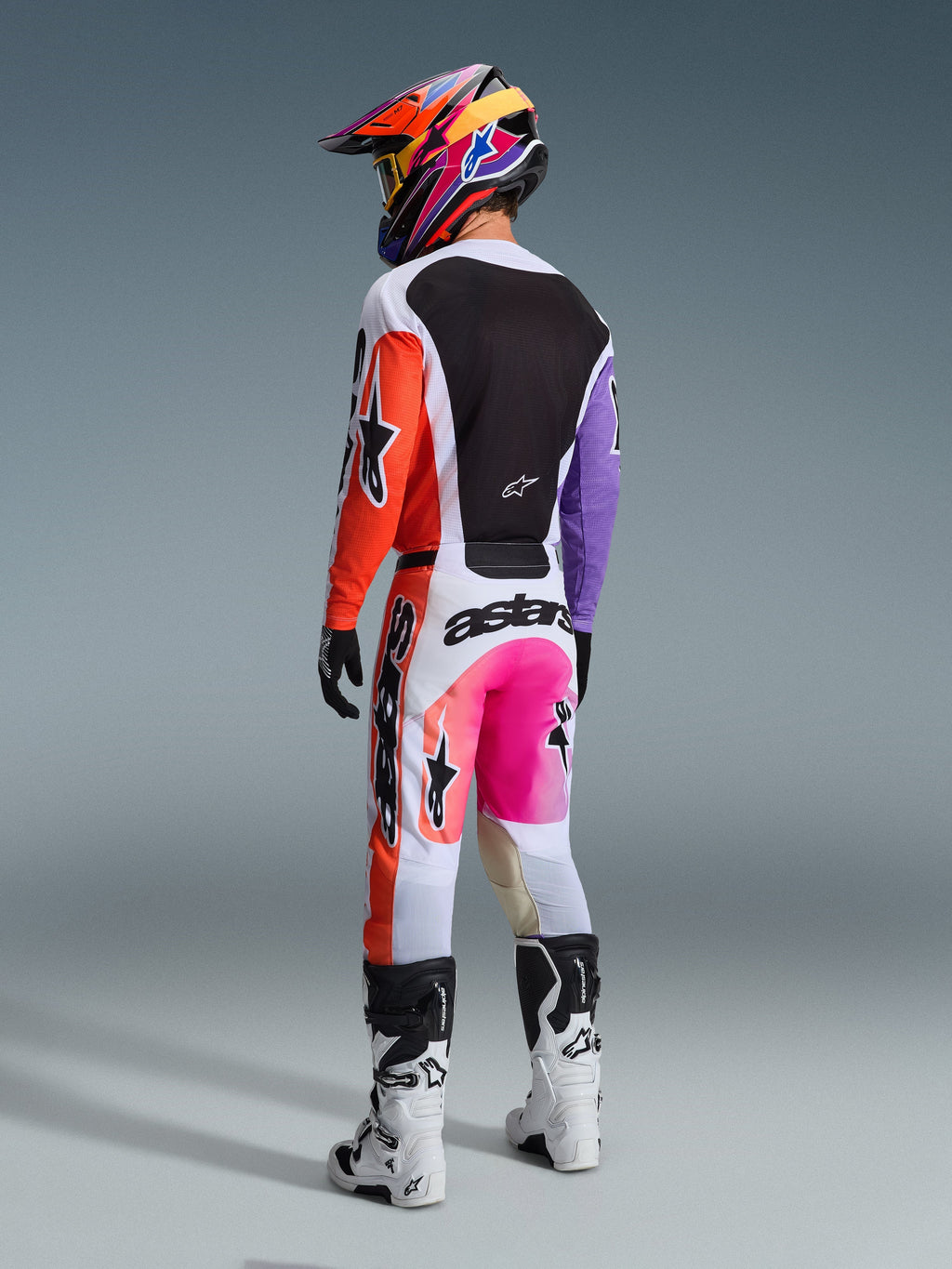 Motocross rider from behind wearing the Alpinestars 2026 Racer Air Portl Pants in Multicolor, featuring white, orange, and purple panels with neon pink accents and bold black branding, paired with a matching jersey, colorful patterned helmet, and white and black boots.