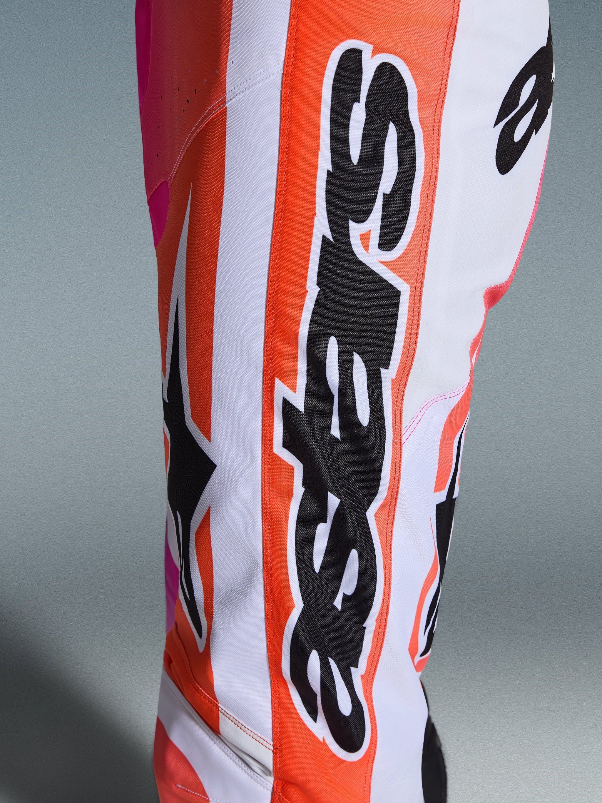 Alpinestars 2026 Racer Air Portl Pants, Motocross Pants, Orange White and Pink, close-up showing large black Alpinestars branding and star logo, perforated fabric for ventilation, durable off-road racing apparel