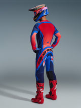 Motocross rider from behind wearing the Alpinestars 2026 Supertech Vista Jersey in Dark Navy, Hot Coral, and Cobalt, paired with matching motocross pants, red boots, and a colorful helmet, coordinated professional racing gear
