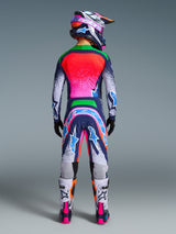 Motocross rider seen from the back wearing the Alpinestars Limited Edition Supertech Records Gear Set, vibrant multicolor jersey and pants with pink, orange, and blue splatter design, matching helmet, black gloves, and white and pink boots, professional off-road racing gear.
