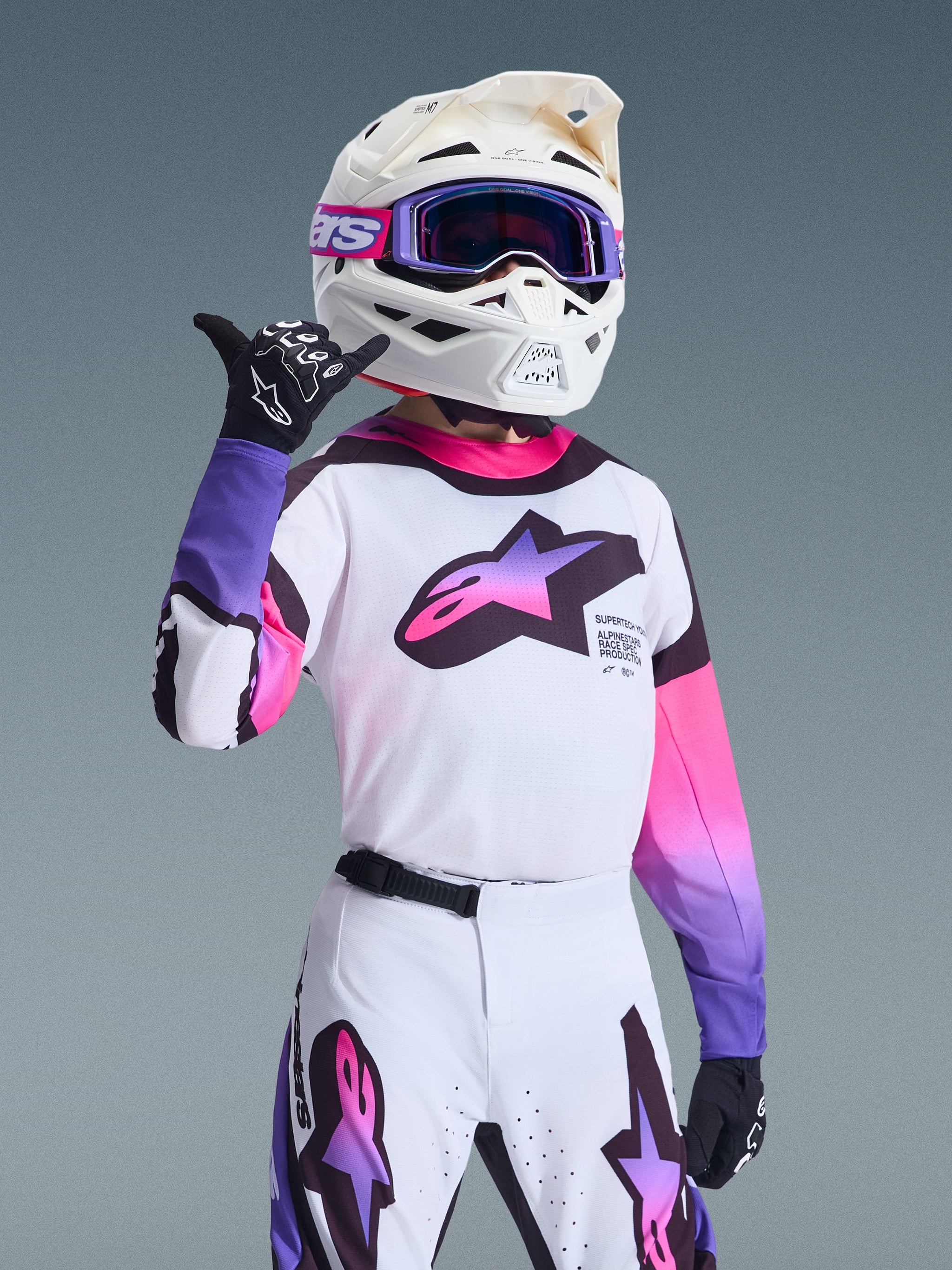 Youth motocross rider wearing Alpinestars 2026 Youth Supertech Vista Jersey, White with Purple and Black accents, featuring a gradient chest logo, matching white helmet, and purple goggles, professional racing outfit for off-road performance