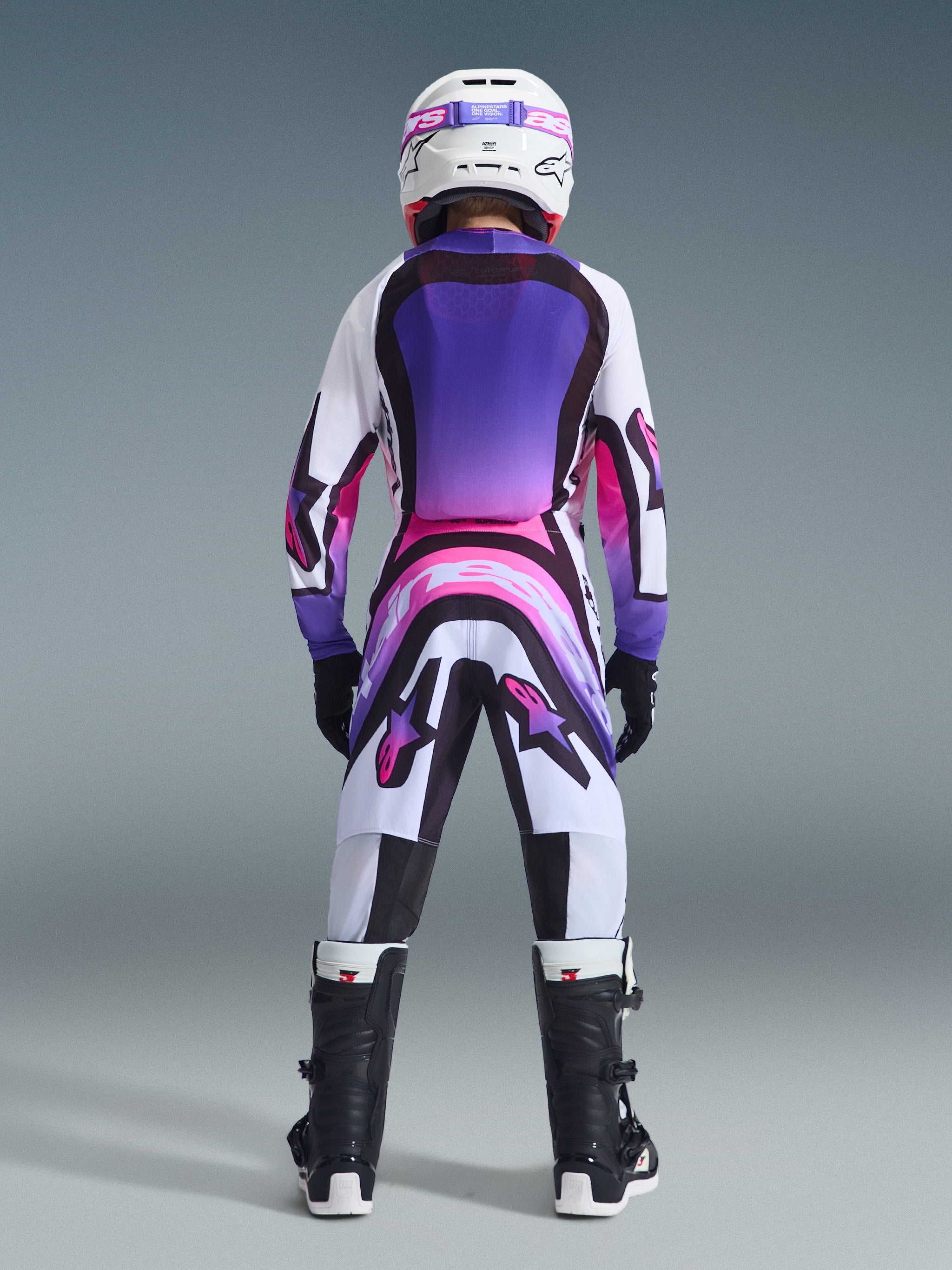 Rear view of a young motocross rider wearing the Alpinestars 2026 Youth Supertech Vista Jersey, featuring a white base with purple and pink gradient panels, paired with matching pants, white helmet, and black boots for a coordinated off-road racing look.