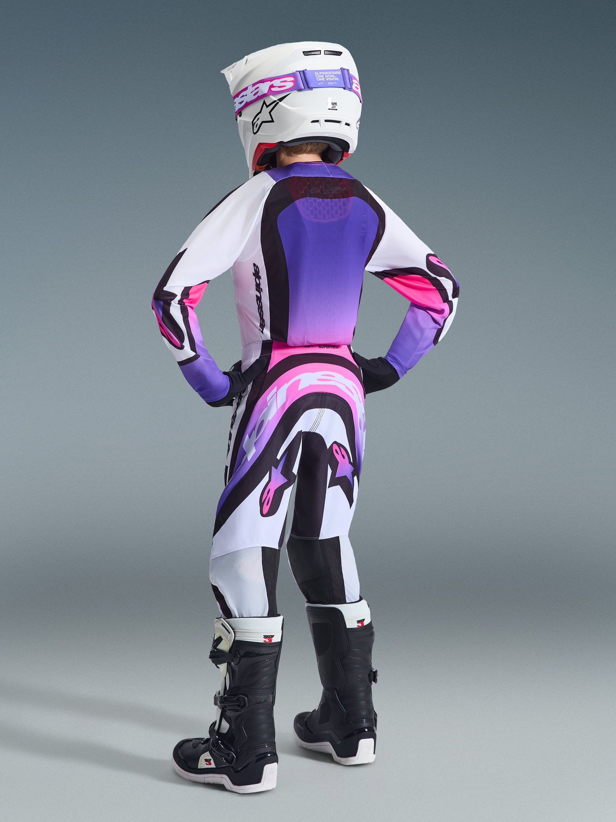 Youth motocross rider wearing complete racing gear, White Purple and Black 2026 Supertech Vista Jersey, matching motocross pants with pink and purple accents, black boots, white helmet with purple goggles, rear view of coordinated professional outfit for off-road racing