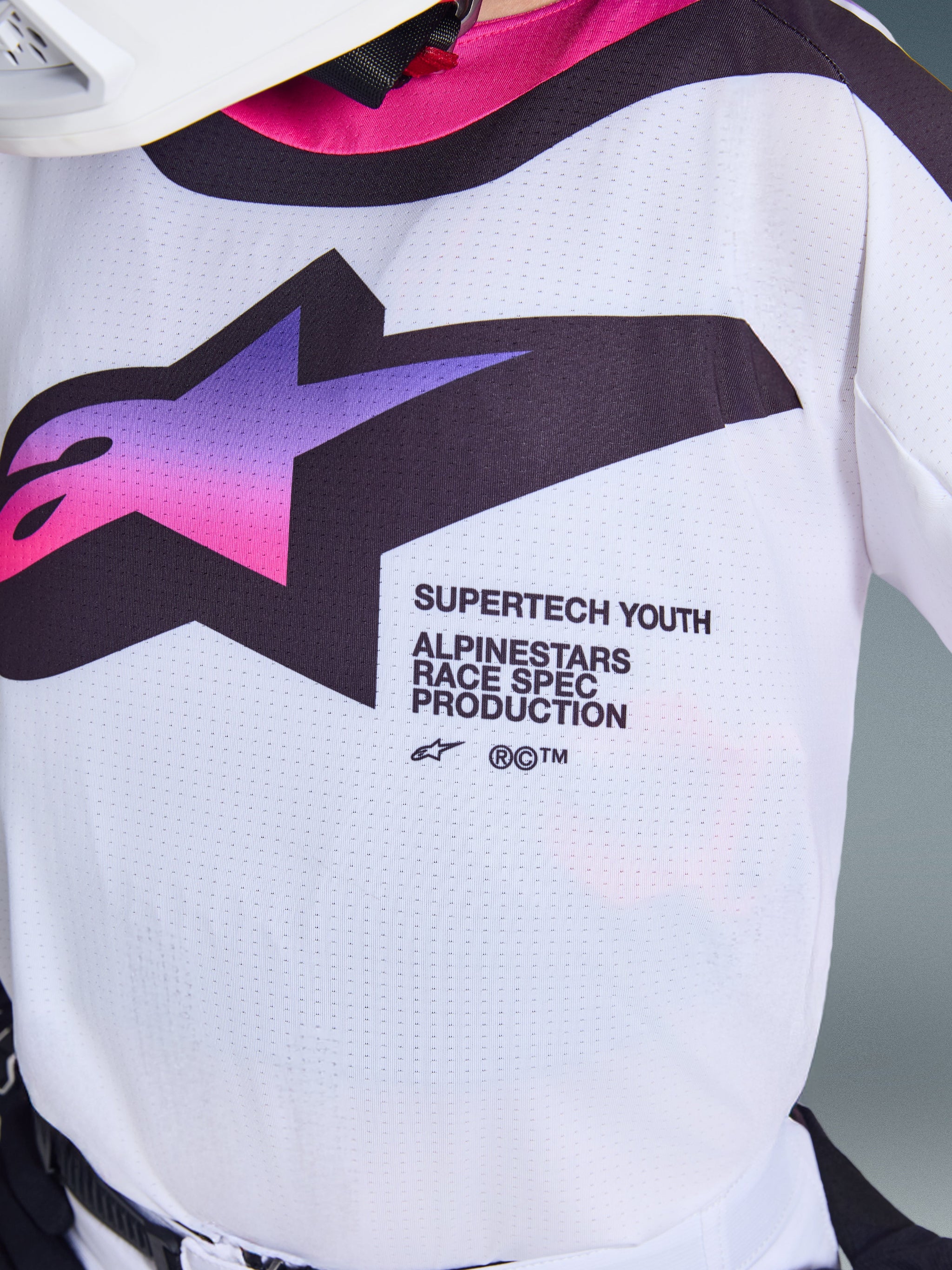 Alpinestars 2026 Youth Supertech Vista Jersey, Motocross Jersey, White with Purple and Pink gradient A-star logo, close-up of perforated stretch mesh fabric and magenta collar, featuring Supertech Youth Race Spec branding for off-road performance.