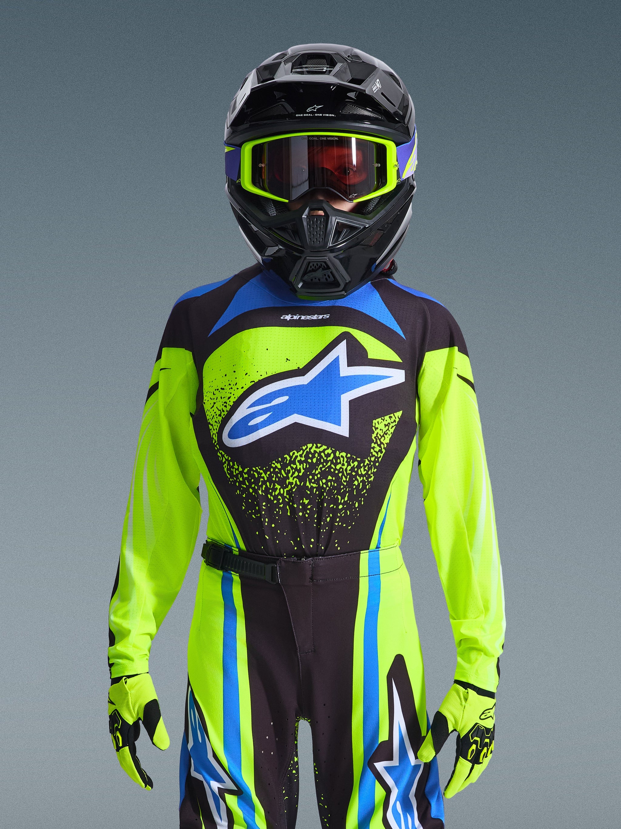 Alpinestars 2026 Youth Supertech Nomur Jersey, Yellow Fluo with Dark Navy and Blue accents, worn by a rider with a black motocross helmet, fluo yellow goggles, and matching gloves, featuring a bold blue star logo on the chest, pro-level off-road racing gear