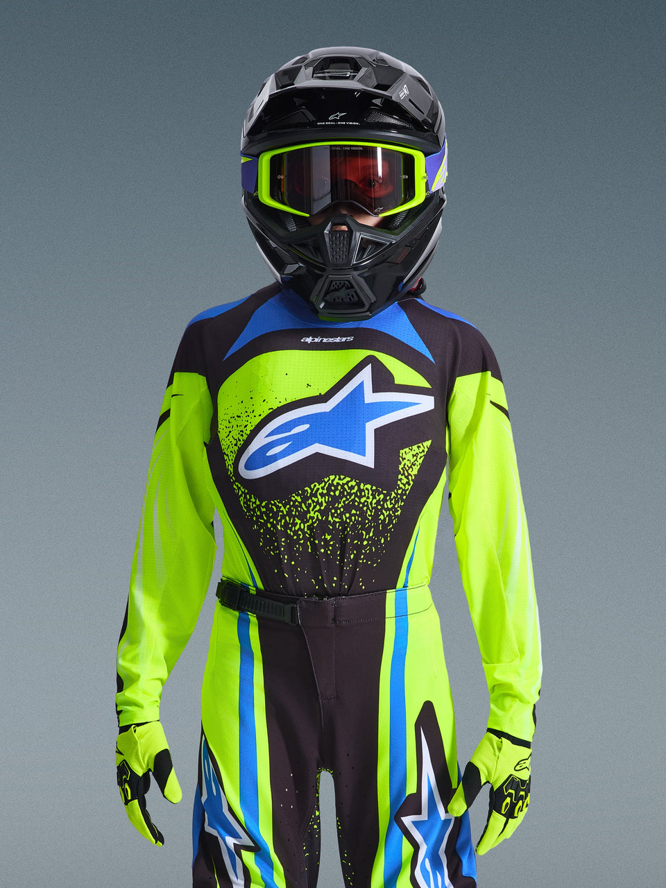 Alpinestars 2026 Youth Supertech Nomur Jersey, Yellow Fluo with Dark Navy and Blue accents, worn by a rider with a black motocross helmet, fluo yellow goggles, and matching gloves, featuring a bold blue star logo on the chest, pro-level off-road racing gear