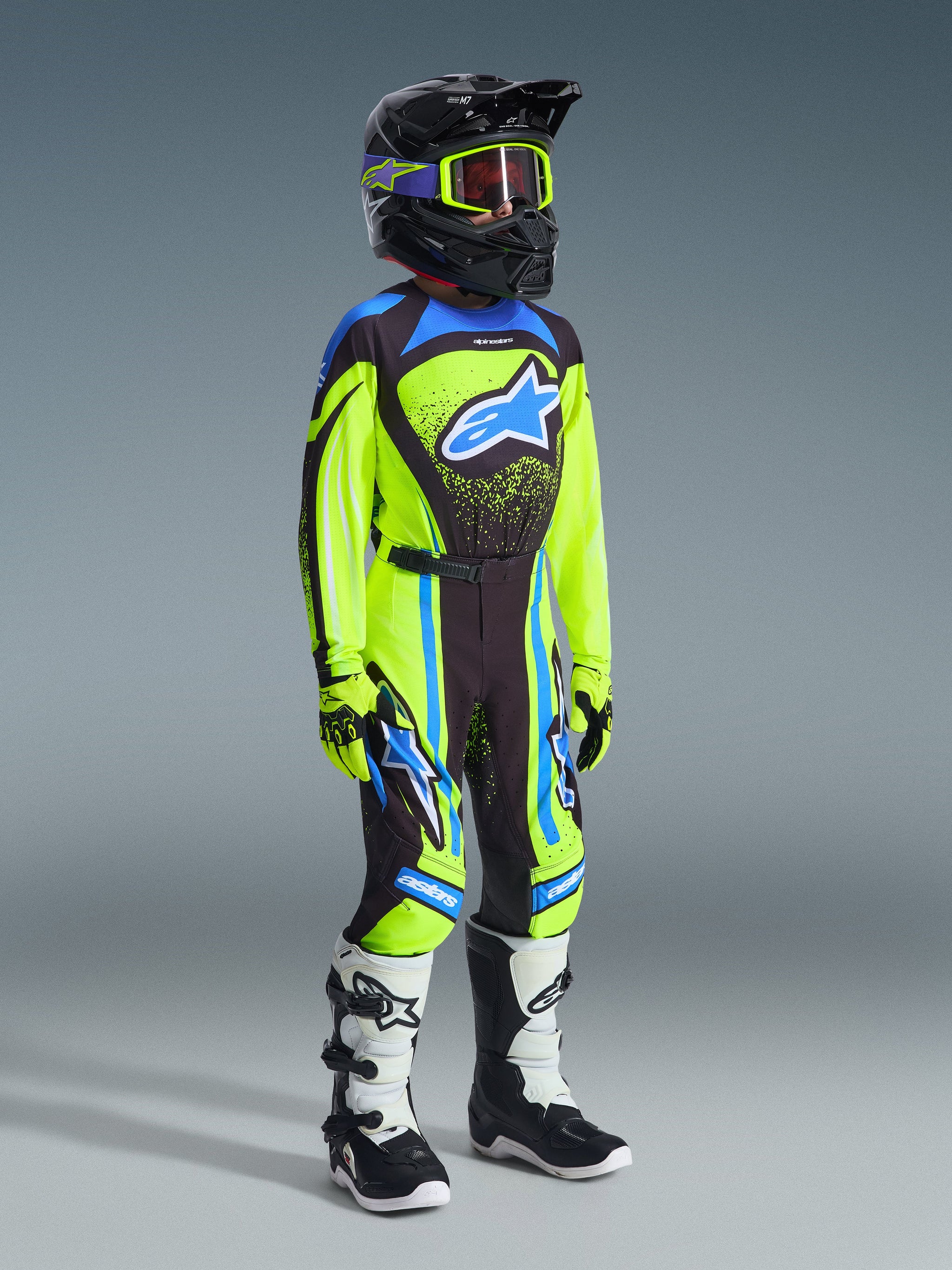 Youth motocross rider wearing complete racing gear, 2026 Youth Supertech Nomur Jersey in Yellow Fluo and Dark Navy, matching pants with blue accents, black helmet with fluorescent yellow goggles, white and black boots, coordinated outfit for off-road racing