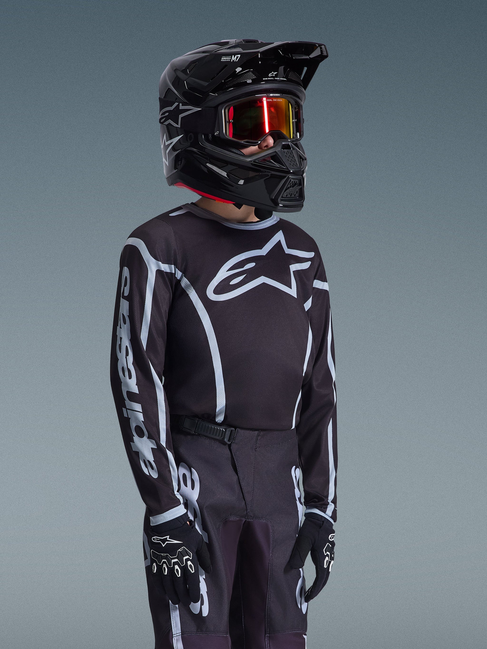 Alpinestars 2026 Youth Fluid Apex Jersey in Black and Gray, worn by a young rider with a matching black helmet and tinted goggles, featuring prominent Alpinestars branding on the chest and sleeves, part of a coordinated motocross gear set.