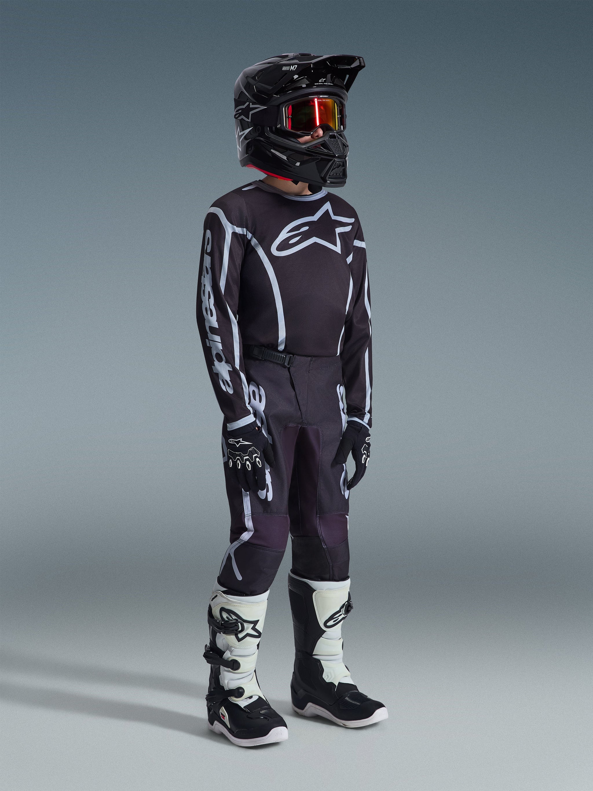 Youth motocross rider wearing the 2026 Youth Fluid Apex Jersey in Black Gray, matching black and gray pants with white graphics, black helmet with red-tinted goggles, black gloves, and white and black motocross boots, complete off-road racing kit.