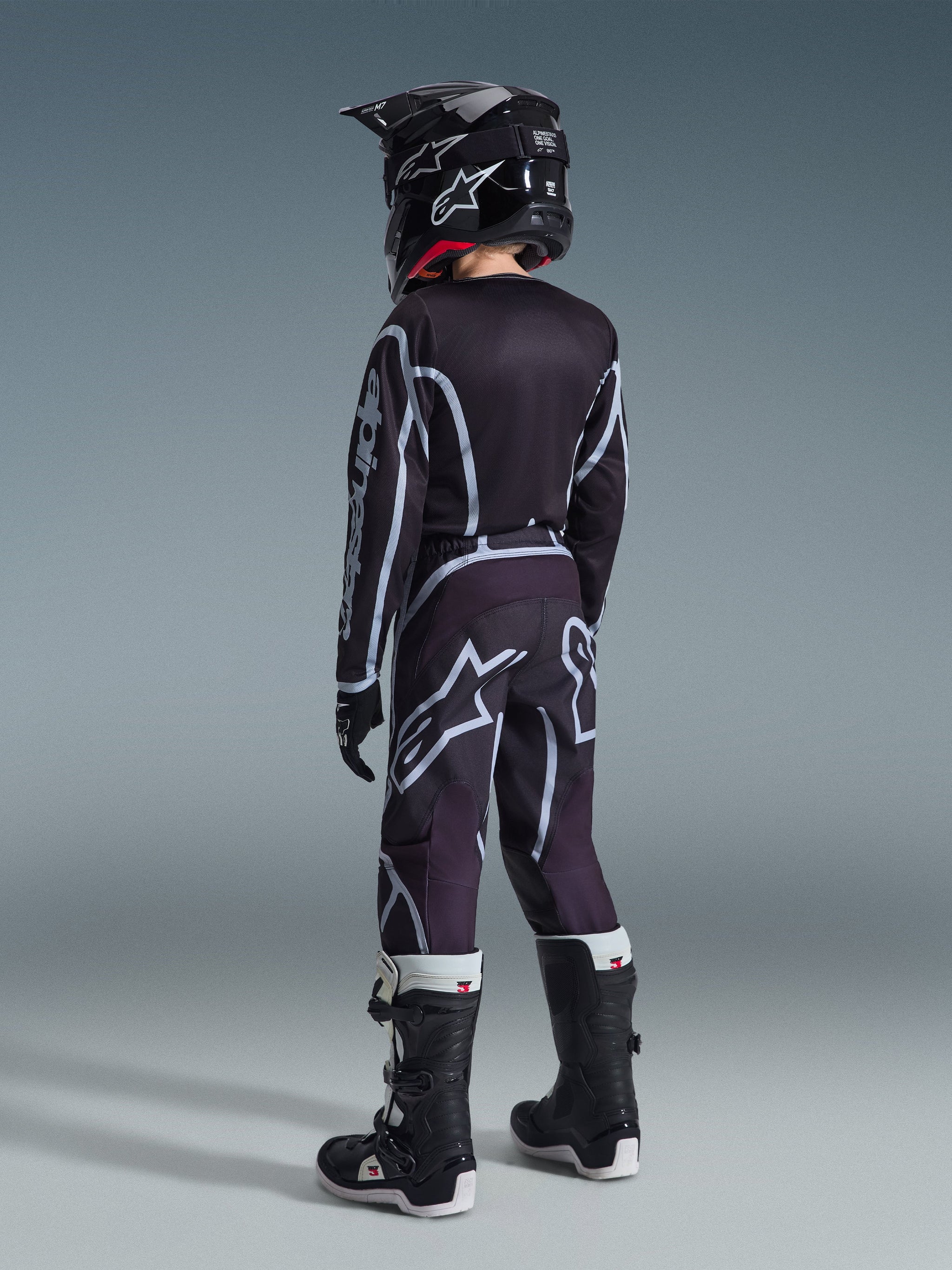 Alpinestars 2026 Youth Fluid Apex Jersey, Motocross Jersey, Black and Gray, worn with matching Youth Fluid Grid Pants, black helmet, and black and white boots, back view showing full off-road racing gear for young riders