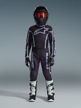 Young motocross rider wearing the Alpinestars 2026 Youth Fluid Apex Jersey in Black and Gray, coordinated with matching pants, black helmet with red iridescent goggles, black and white gloves, and motocross boots, complete gear set for youth off-road racing