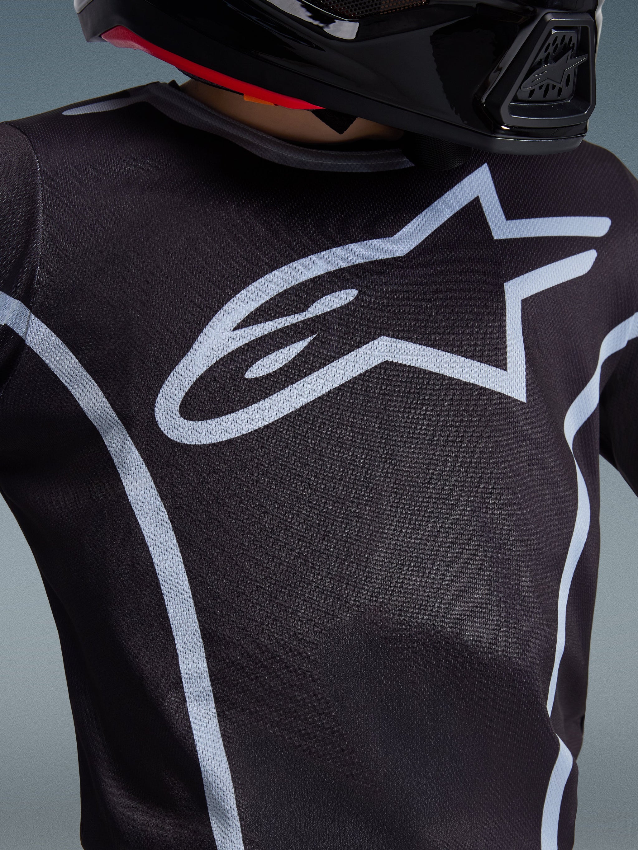 Alpinestars 2026 Youth Fluid Apex Jersey, Close-up of chest design, Black and Gray, featuring a prominent light gray star logo and athletic piping, worn with a black helmet, performance motocross gear for youth riders