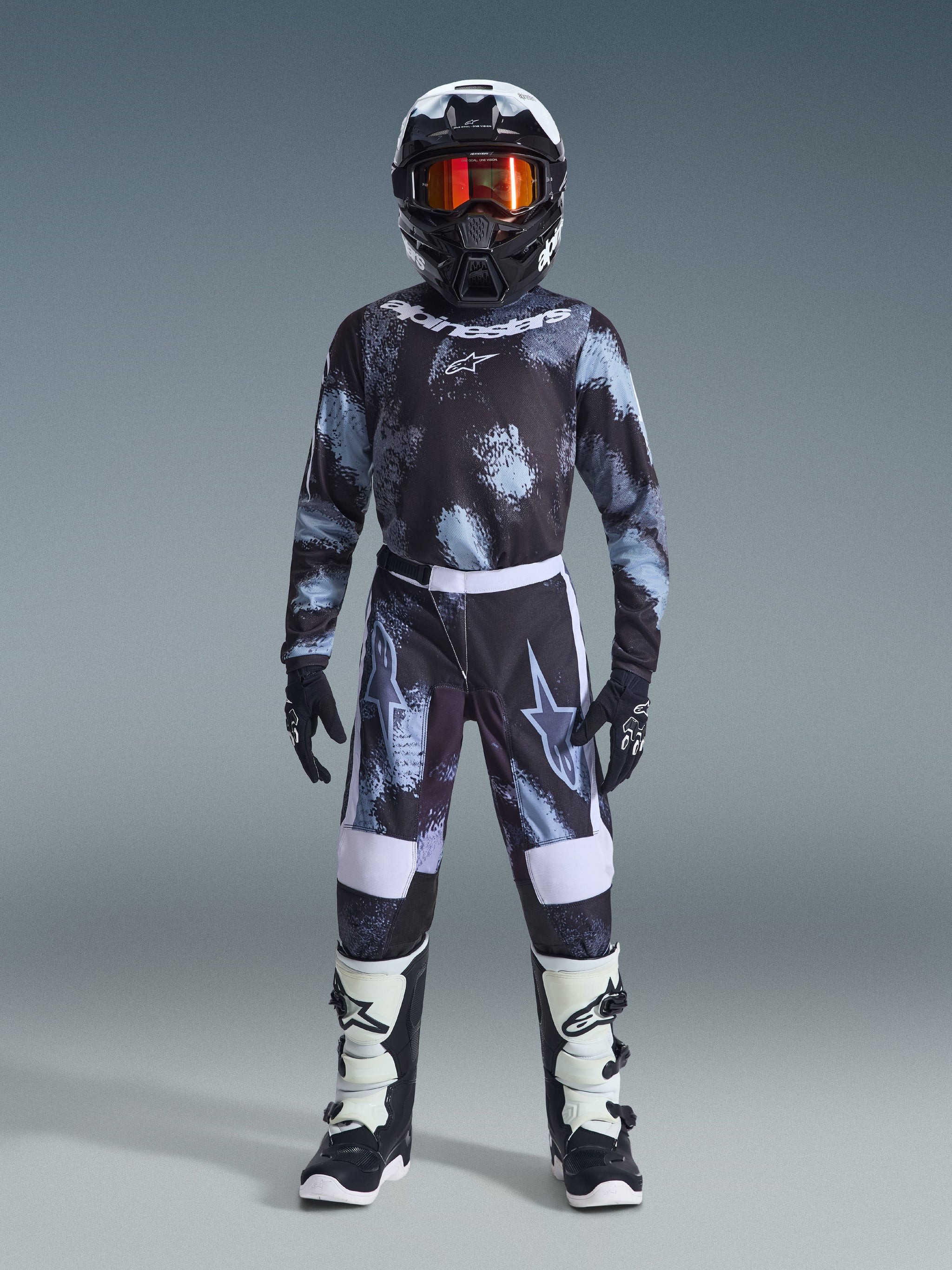 Youth motocross rider wearing complete racing gear, Alpinestars 2026 Youth Fluid Lahnd Jersey in Iron Camo with black, grey, and light blue pattern, matching pants, black and white helmet with orange tinted goggles, black and white boots, coordinated gear set for off-road riding