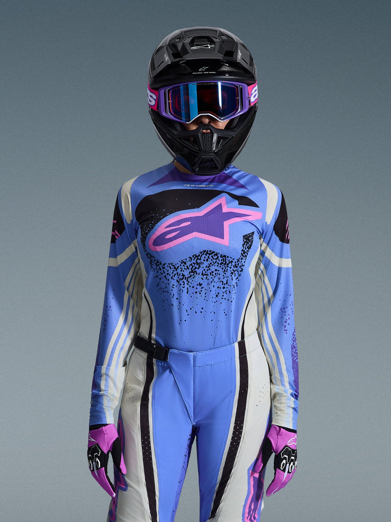 Woman motocross rider wearing the Alpinestars 2026 Stella Techstar Nomur Jersey in Light Blue and Off White, featuring black graphic patterns and a pink logo, styled with matching pants, black helmet, purple goggles, and pink gloves for a coordinated off-road racing look.