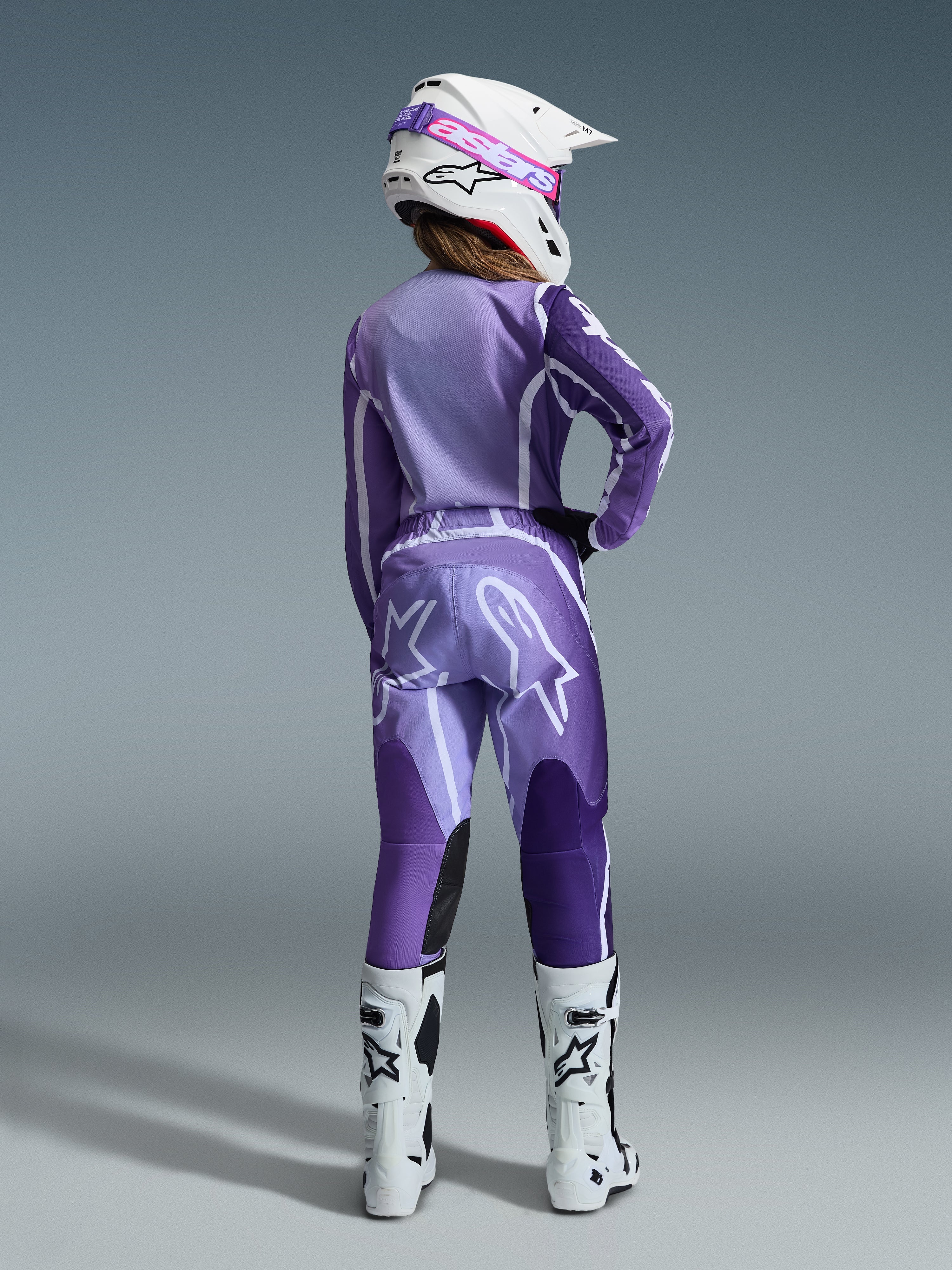 Alpinestars 2026 Stella Fluid Apex Gear Set, Women's Motocross Gear, Purple and Lavender with white accents, female rider seen from the back wearing matching jersey and pants, white helmet with pink goggles, and white boots, coordinated athletic racing outfit