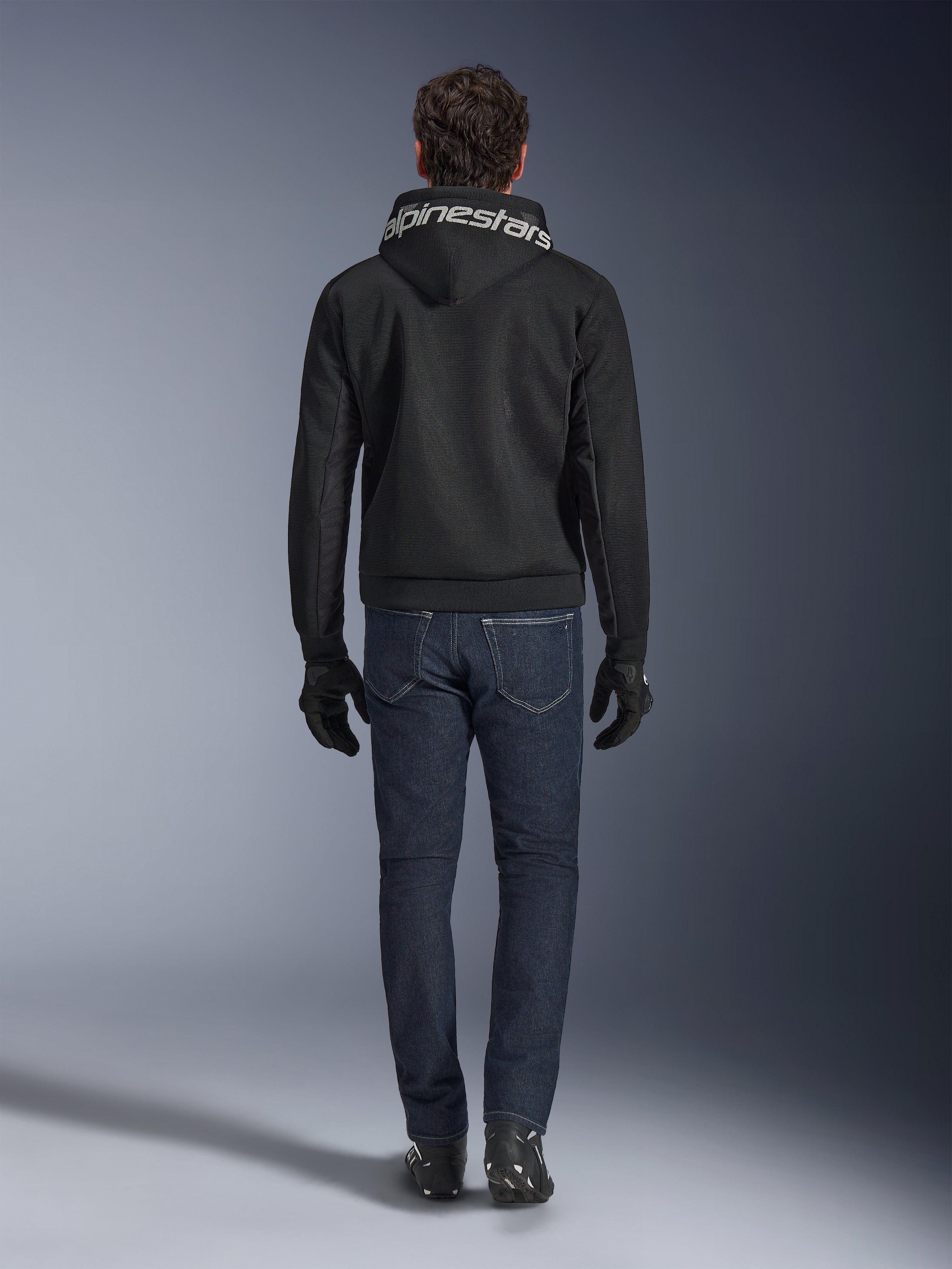 Alpinestars Chrome Superairflow Hoodie, Black motorcycle hoodie with white branding on the hood, viewed from the back, worn with dark blue jeans, black gloves, and black and white boots, urban sport riding gear