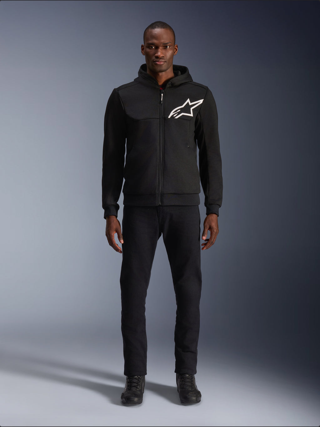 Alpinestars Chrome V2 Sport Hoodie, Black and White motorcycle hoodie, featuring a large white Alpinestars logo on the chest, zip-up front and hood, worn by a model standing with black pants and boots, urban-style protective riding gear.