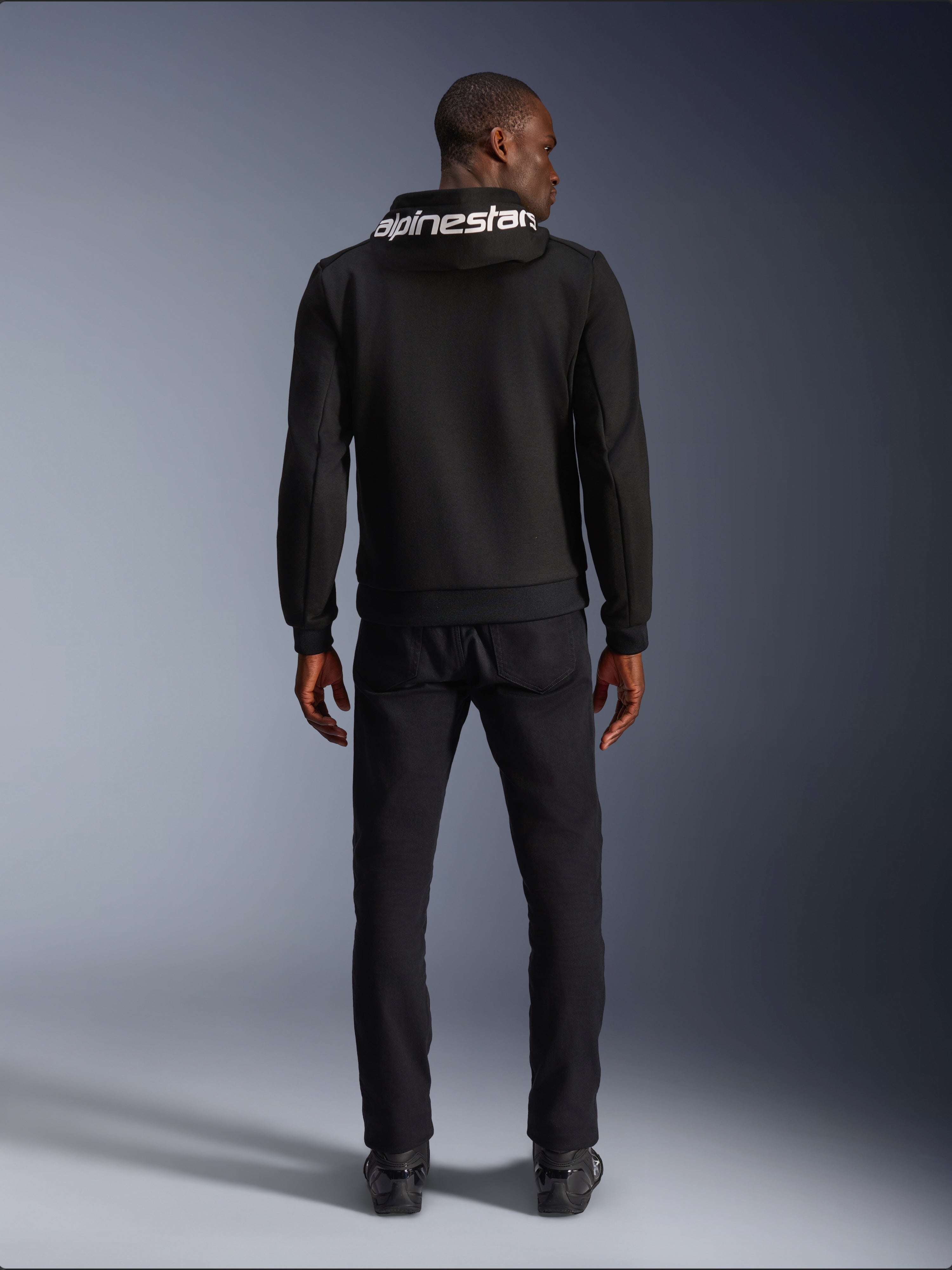 Alpinestars Chrome V2 Sport Hoodie, Motorcycle Hoodie, Black, back view of model wearing black tech fleece hoodie with white Alpinestars logo on the hood, paired with black pants and boots, casual urban riding style