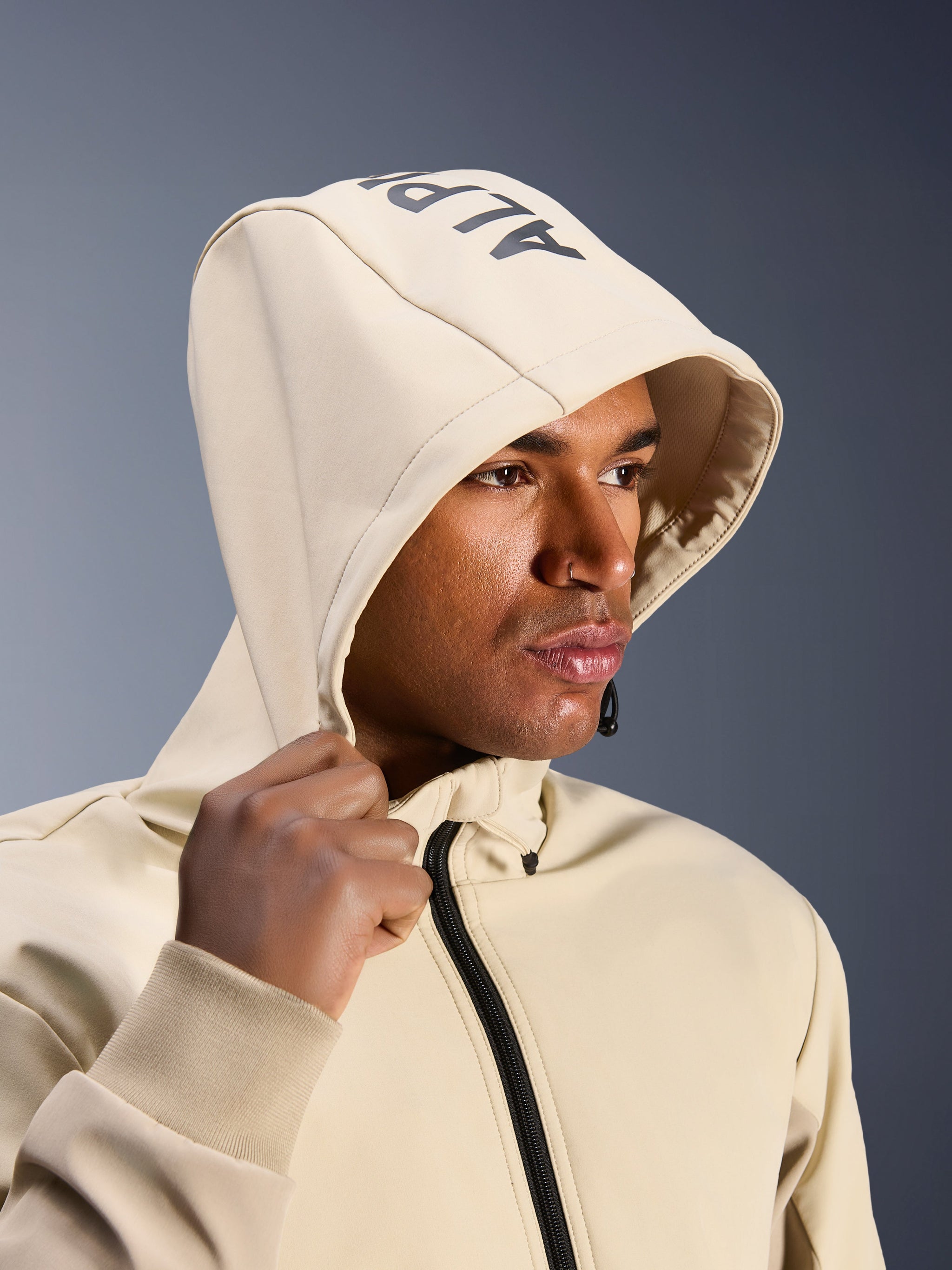 Alpinestars Radium Tech Hoodie in Mastic Aluminum, close-up of a man wearing the hood up, featuring Alpinestars branding on the top, urban motorcycle jacket with technical windproof shell, modern streetwear aesthetic for city riding
