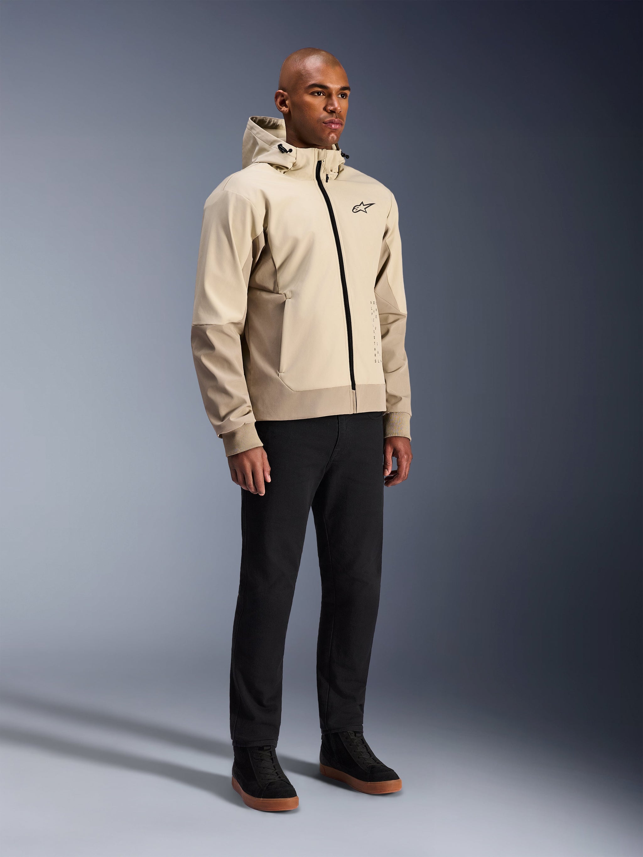 Alpinestars Radium Tech Hoodie, urban motorcycle jacket, Mastic Aluminum light beige, worn with black pants and black high-top sneakers, featuring a technical windproof shell, front zipper, and removable hood, modern streetwear design for urban riding