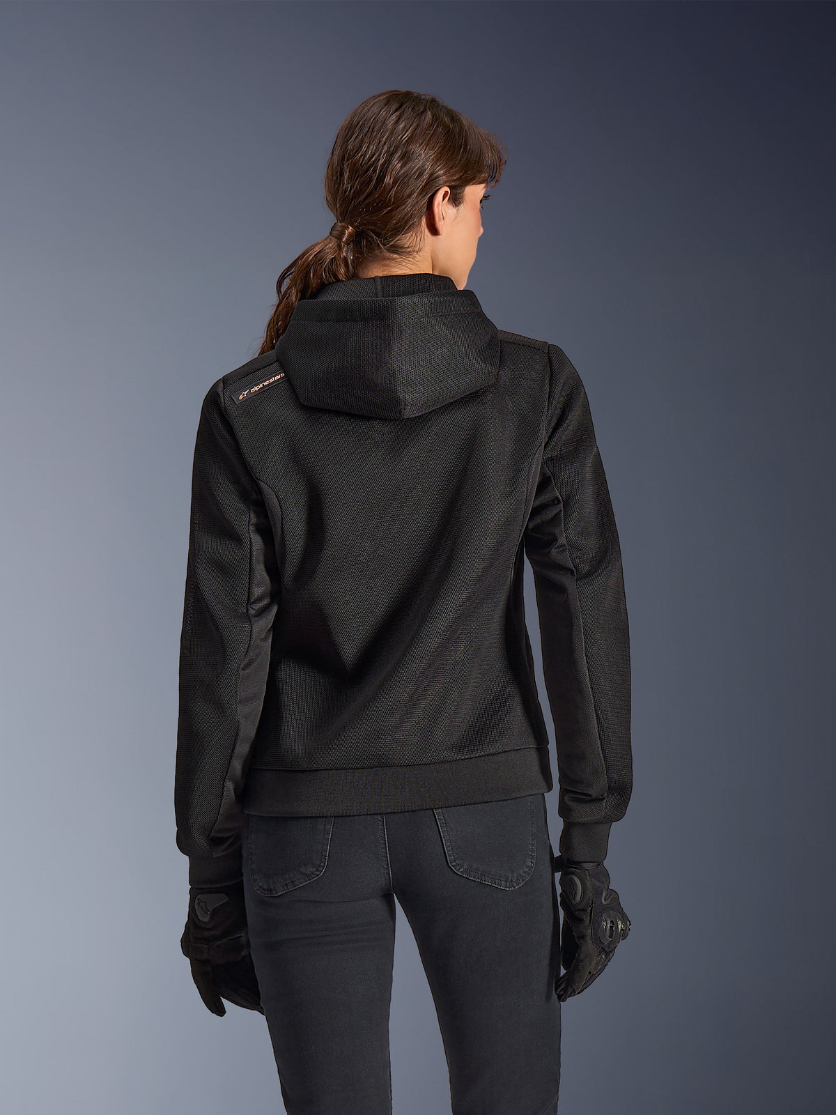 Alpinestars Stella Chrome Superairflow Hoodie, back view of woman wearing black technical 3D mesh motorcycle hoodie, features engineered hood and copper logo on shoulder, worn with black motorcycle gloves and pants.