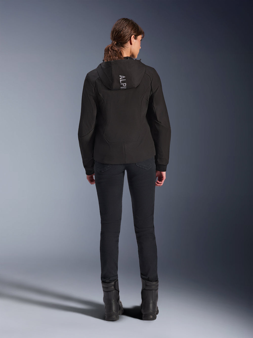 Alpinestars Woman Stella Radium Tech Hoodie, Black, Rear view of model wearing lightweight 3-layer windproof tech hoodie with Alpinestars logo on the hood, paired with black pants and boots, urban motorcycle riding apparel