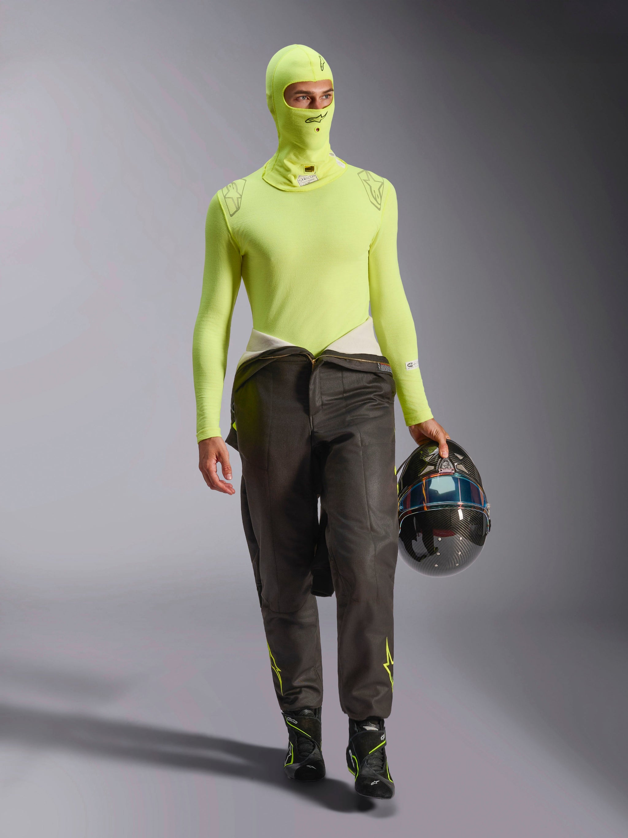 Male racing driver wearing Alpinestars ZX Evo V3 Balaclava in Yellow Fluo, matching long-sleeve base layer top, dark racing pants, and black racing boots, holding a carbon fiber helmet, professional automotive racing gear