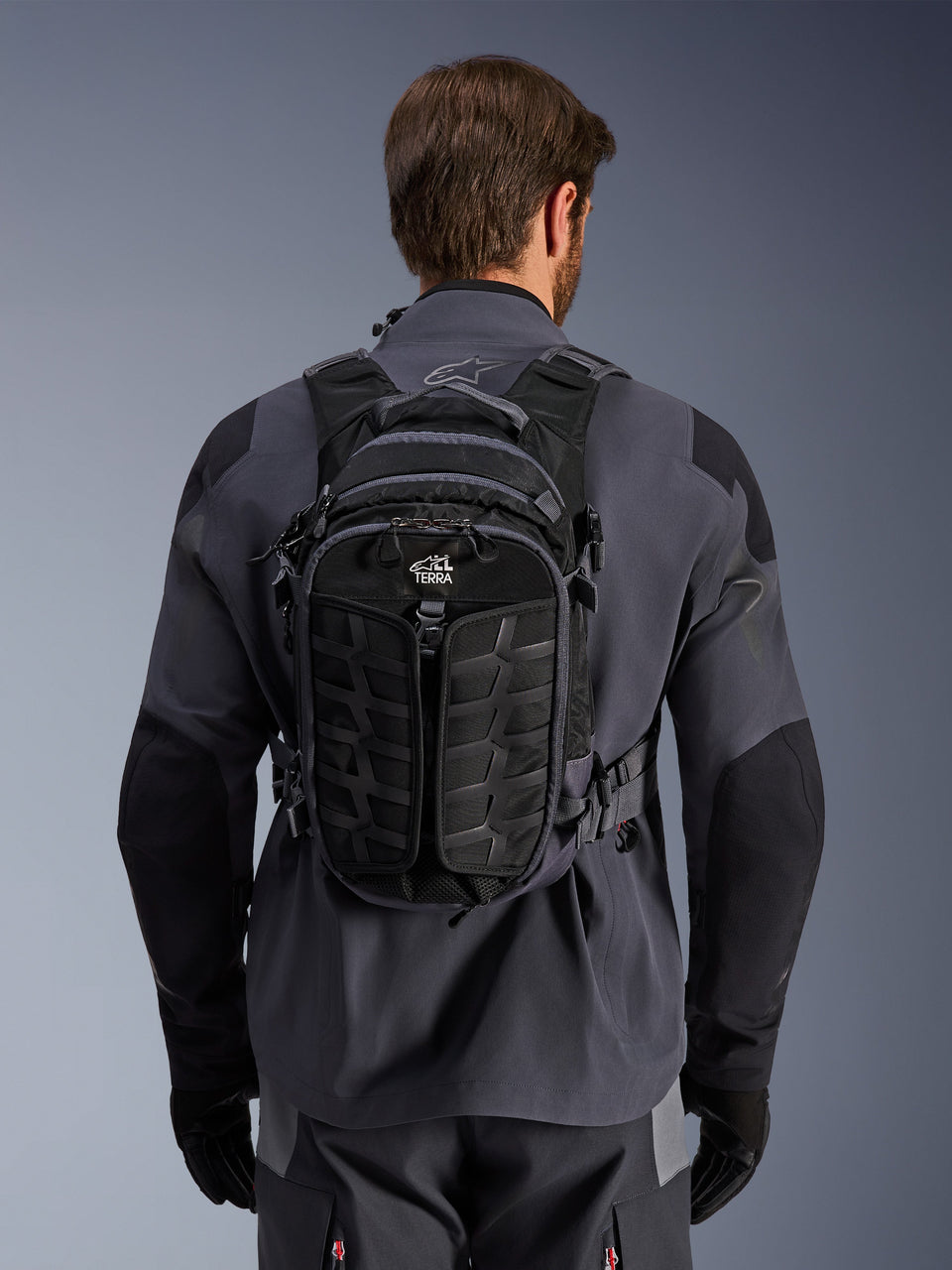 Alpinestars AT-15 Back Pack, adventure motorcycle backpack, Black Deep Gray, worn by a person over a gray technical riding jacket, showing the back view with 'ALL TERRA' branding, ergonomic design with reinforced textured panels and secure straps.