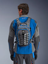 Alpinestars AT-15 Back Pack, adventure motorcycle backpack, Dark Gray and Deep Gray, worn by a rider over a blue and gray jacket, featuring All Terra branding, ergonomic structural design, adjustable straps, and blue zipper accents.